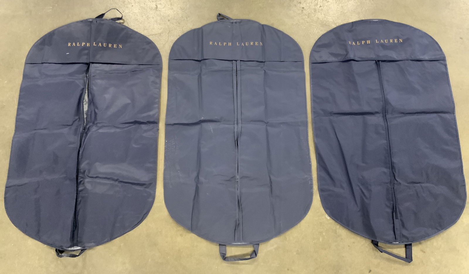 RALPH LAUREN Lot 3 Nylon Garment Bags: Garment bags are trademarked RALPH LAUREN. Each bag is navy toned. Each appears to be nylon. Bags each have handles to hang. Each measures approximately 39 inches tall 24 inches wide. Garment bags, na