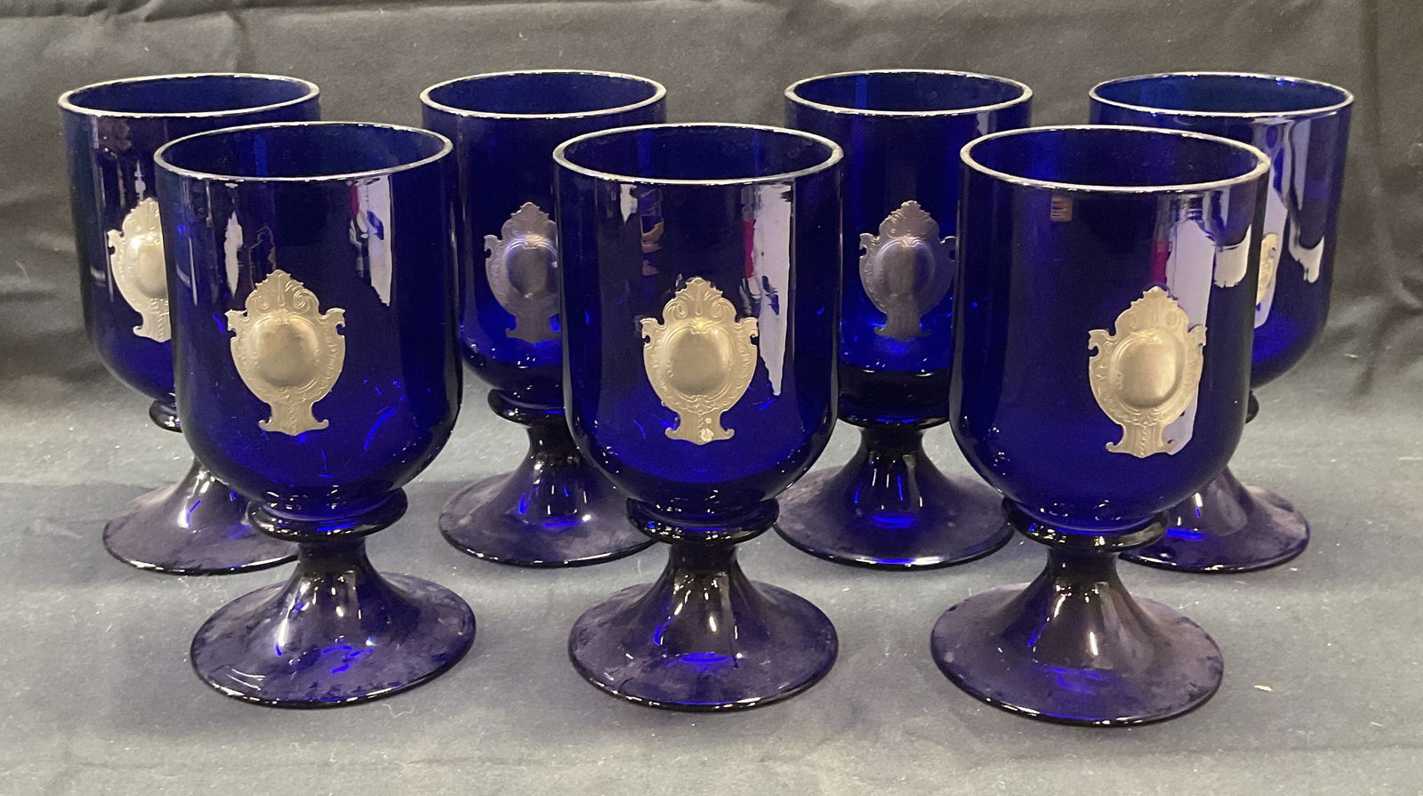 Set 7 Cobalt Blue Glass Goblets w Shield Motif: Set of 7 cobalt blue glass goblets with pewter shield motif decoration. Measure approx. 3 3/8 x 6 3/8 inches each. Pieces have weight. All pieces have some surface wear including scratching observed.