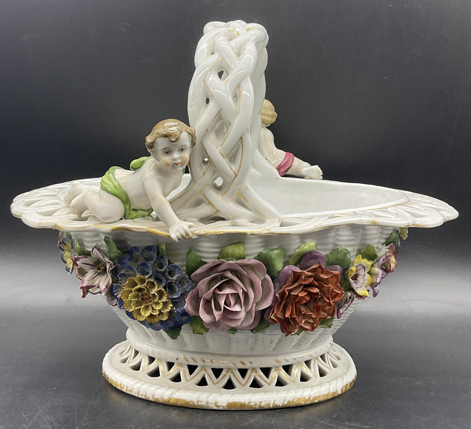 Vintage Trademarked Porcelain Figural Basket: A vintage porcelain basket featuring figurals, raised florals, pierced porcelain, and gilt line work. Multicolored hues throughout the white porcelain basket. Underside trademarked. Measures approx. 1