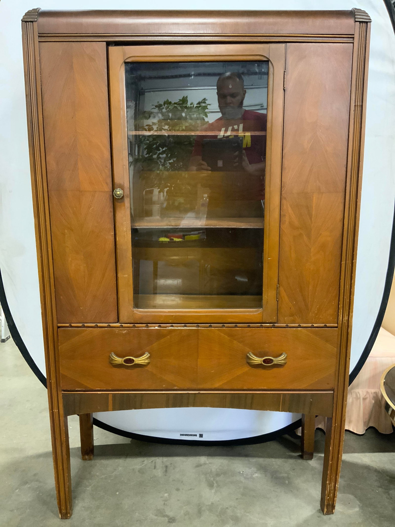 Vintage Wooden China Cabinet W Drawer: China cabinet is tan toned and wooden. Piece has drawer with gilt metal pulls. Top half has windowed door to cabinet Area with 2 shelves. Cabinet handle has floral form. Piece measures approximately 6