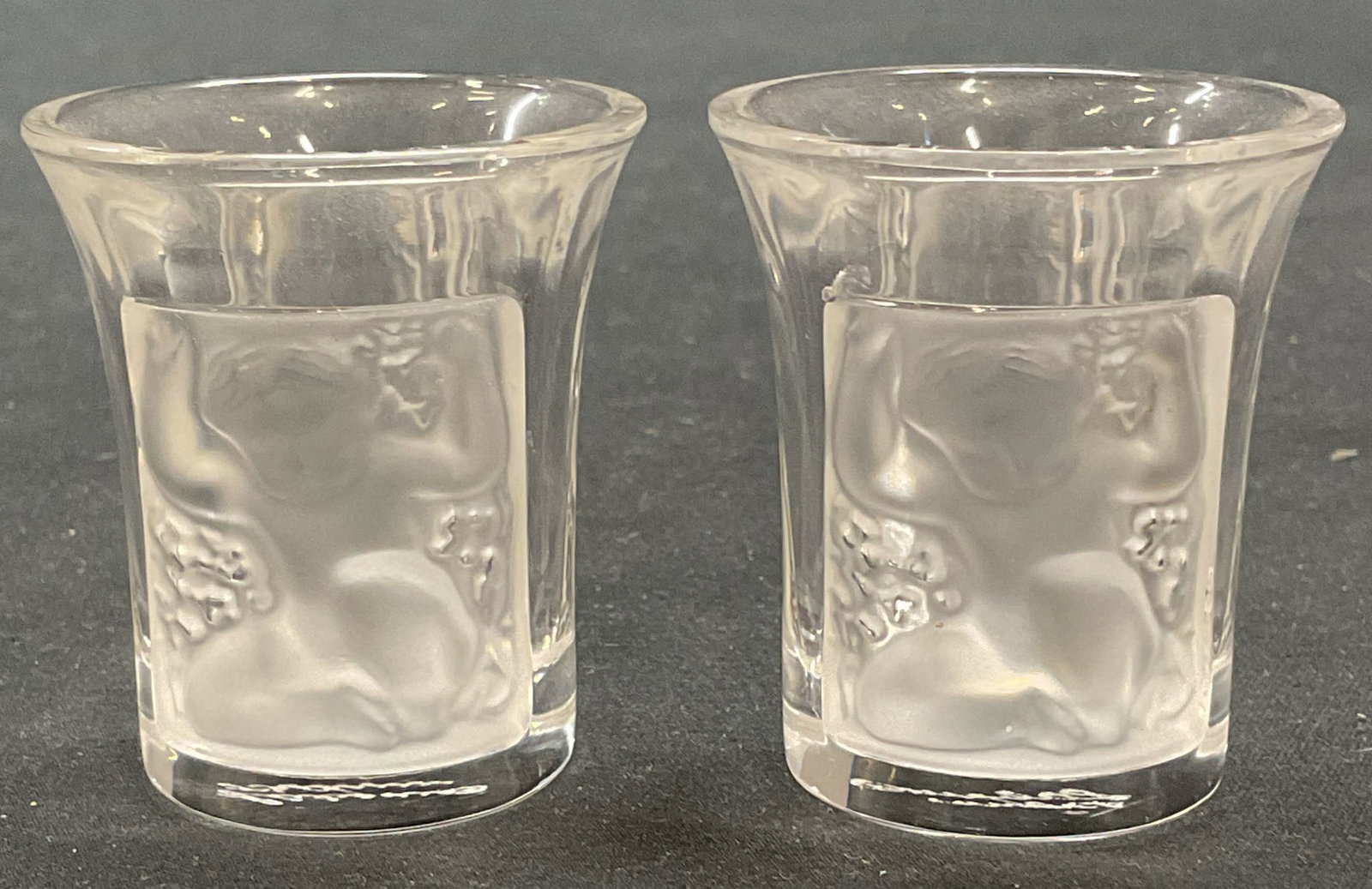 Pair Lalique Crystal Enfants Shot Glasses, France: Pair of Lalique frosted crystal cameo Les Enfants shot glasses. Raised figural baby motif. Signed on undersides. Made in France. Measure approx. 1.5 x 1.75 inches each. Both pieces have some surface w