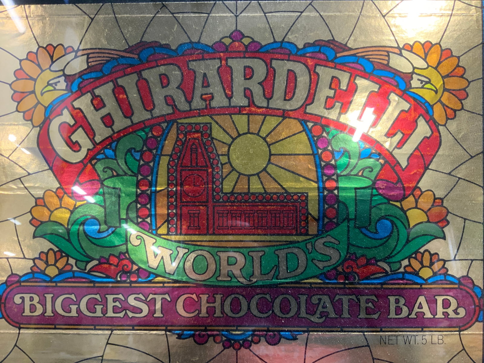 GHIRADELLI World Biggest Choc Bar Wrapper In Frame: Piece reads GHIRADELLI WORLDS BIGGEST CHOCOLATE BAR. Piece is in blue toned metal frame encased in glass. Piece measures approximately 21 inches wide 16.75 inches tall. GHIRADELLI worlds biggest choco