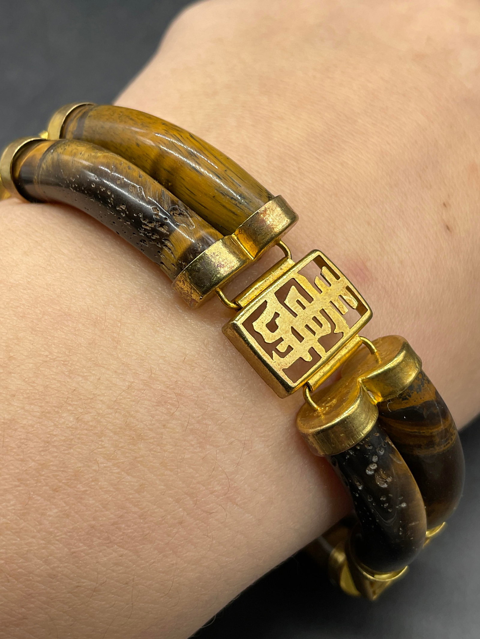 Asian Character Motif Polished Tigers Eye Bracelet: Asian Character Motif Polished Tigers Eye Bracelet. Crafted from gold tone metal and tigers eye. Length measured end to end approx. 8 inches. Box clasp closure. Imperfections to the stones. Surface we