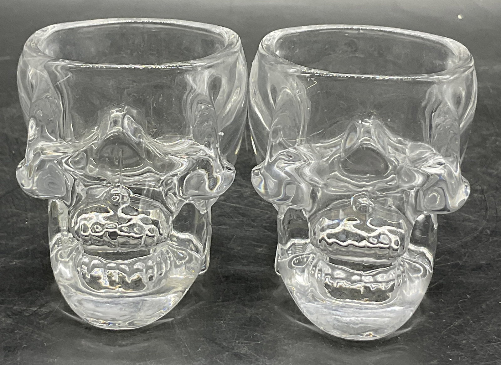 Pair STUDIO CRYSTAL Clear Glass Skull Vessels: A pair of clear glass skull vessels. Stickers to reverse read Studio Crystal by Studio Silversmiths. Measures approx. 2.5H x 3W in. Surface wear consistent with age, use, and exposure. home decor, hom