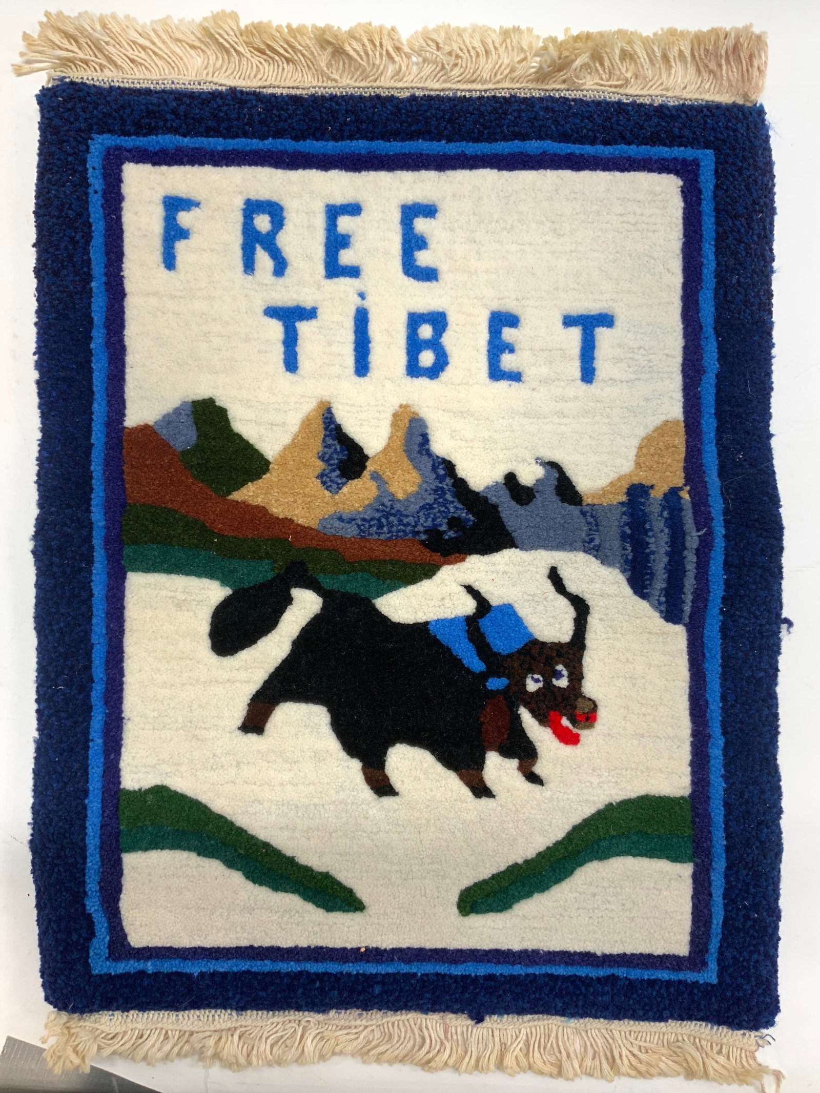 Free Tibet Fringed Wool Rug: Rug appears to be wool, possibly polypropylene/ wool blend. Piece reads FREE TIBET with image of animal and landscape. Piece has cream toned fringes at edges. Rug measures approx 26 inches long 18 inc