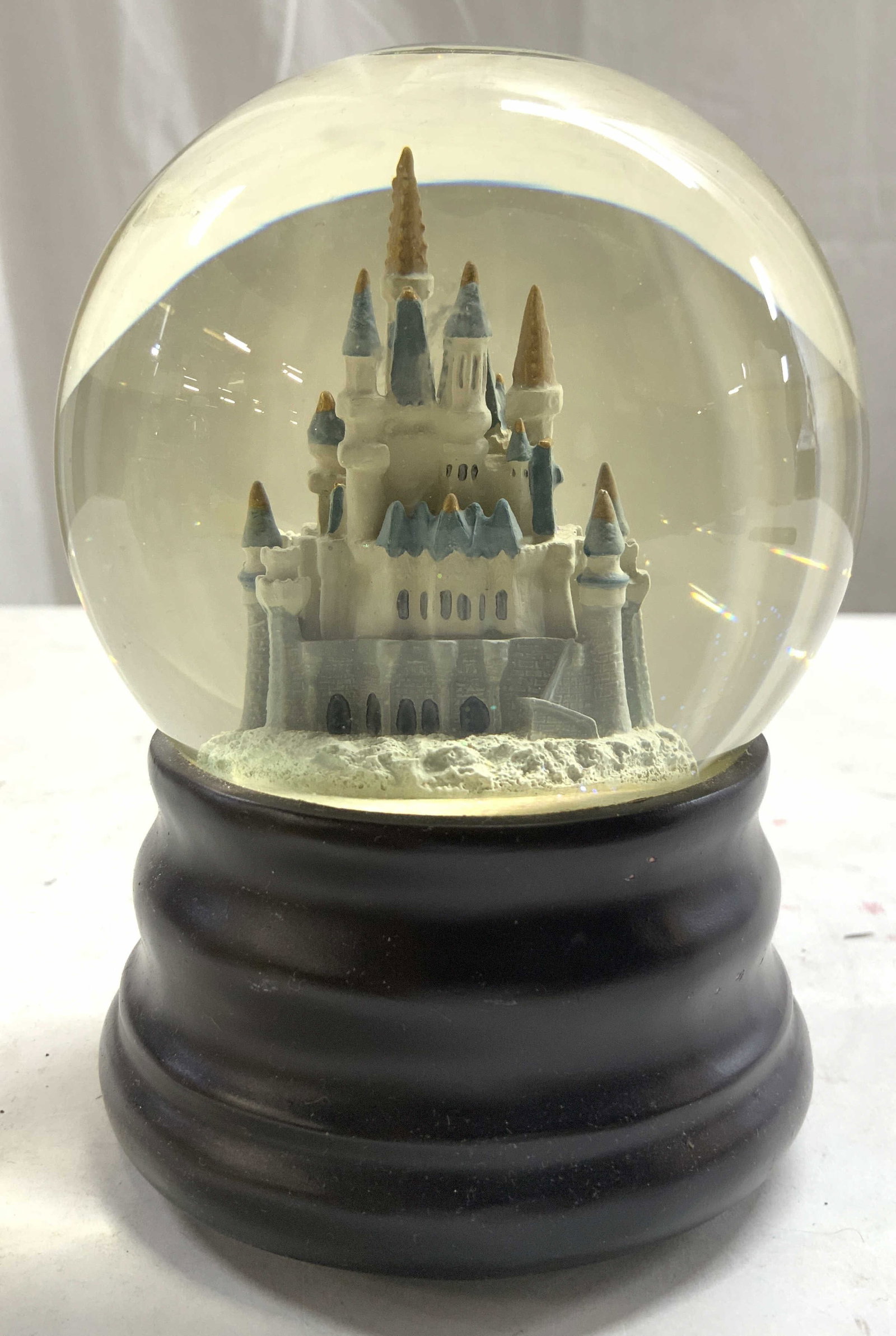 DISNEY Kingdom Snow Globe Music Box: Underside reads DISNEY. Underside also has music box wind with letter S. Snow globe has structure resembling Disney Kingdom inside. Base of piece is brown toned. Measures approximately 5.5 inches tall