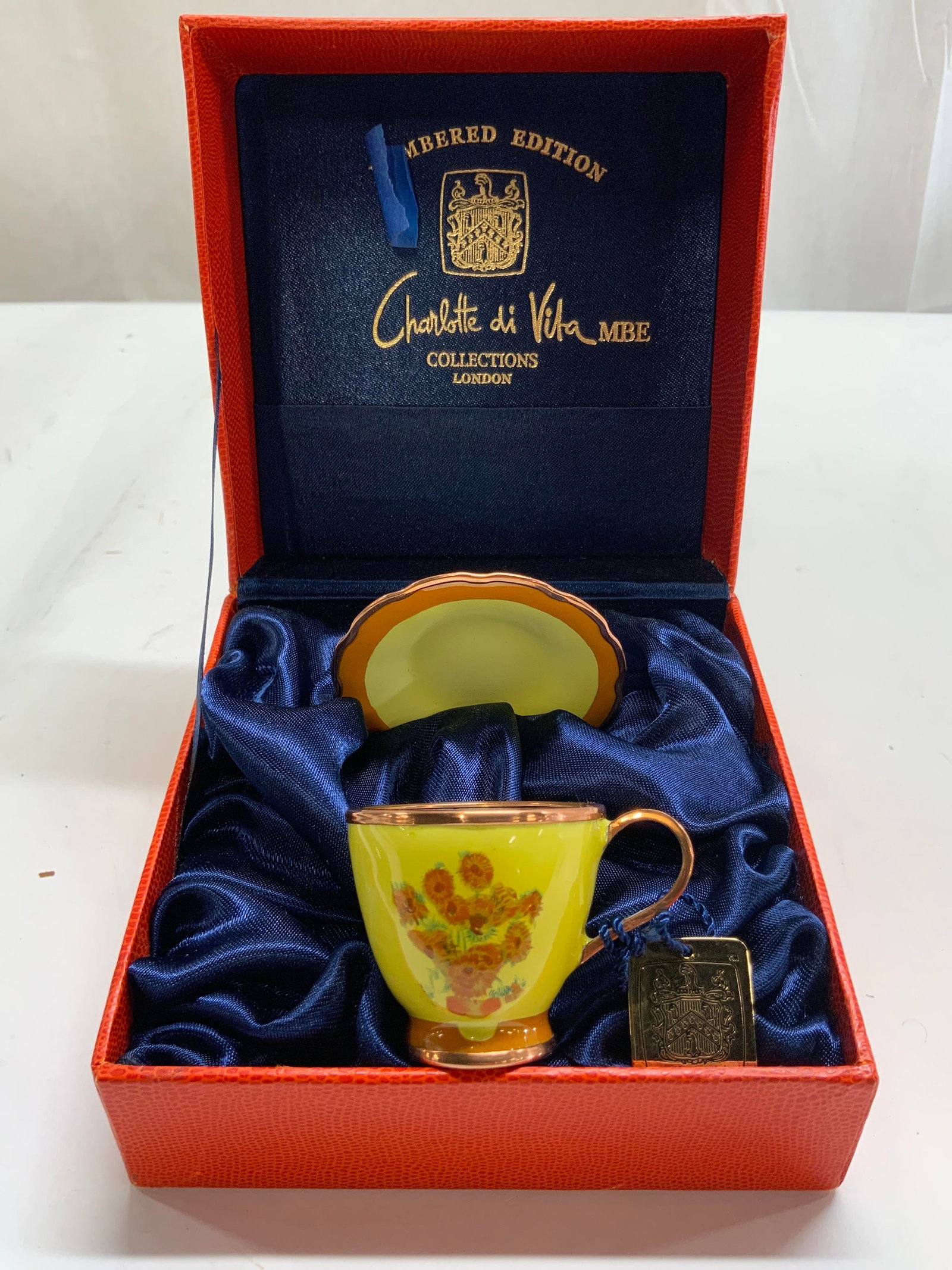 Charlotte Di Vita Mini Porc Cup & Saucer In Box: Underside of lid reads CHARLOTTE DI VITA. Lot includes 1 teacup, 1 saucer, & Original orange toned box. Each piece is yellow and orange toned with floral and leaf detail. Saucer measures approximately