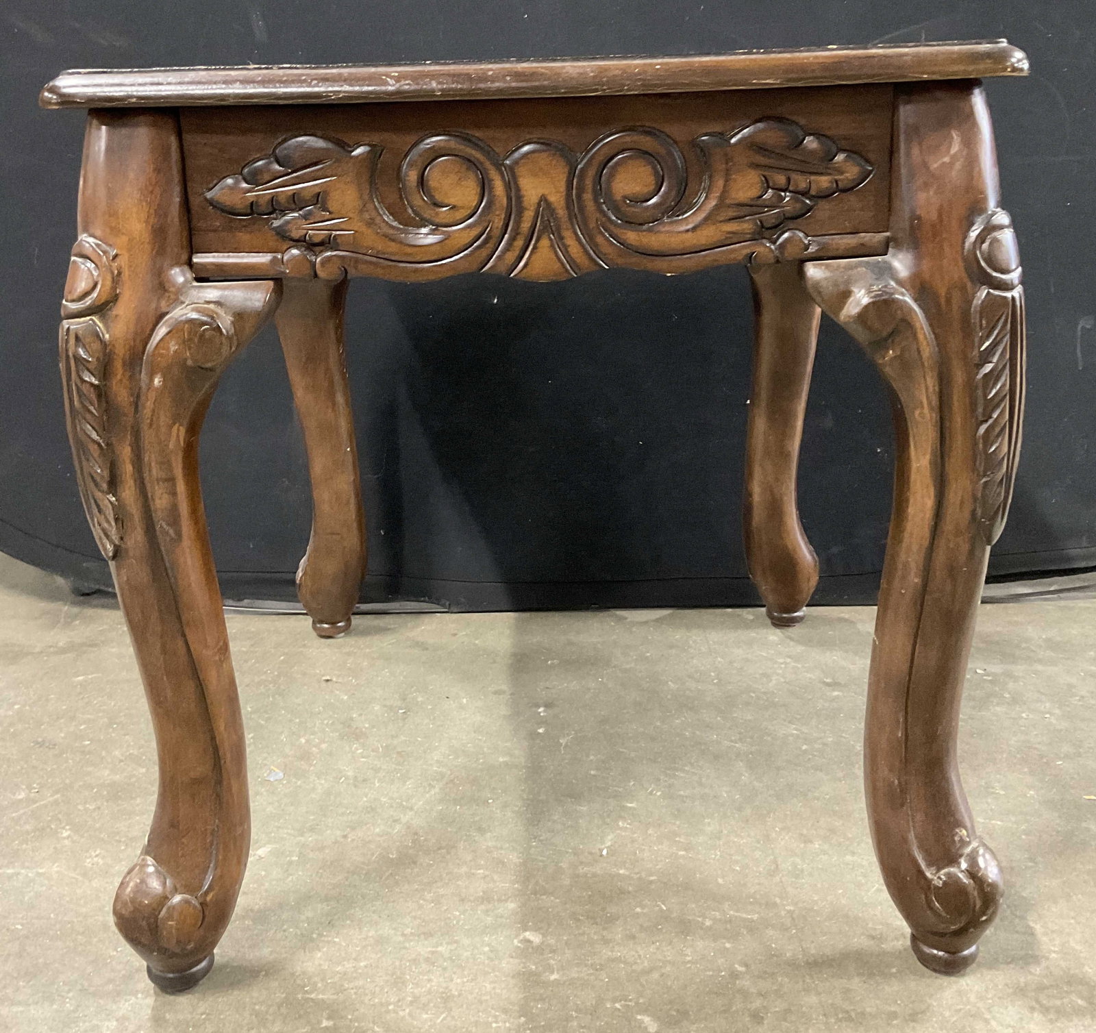 Wooden Side Table W/ Scrolled Feet: A rich brown hued wooden side table with raised woodwork designs and scrolled feet. Measures approx. 21H x 22W x 21D in. Residue, surface wear, and chipping present. Partial stocker to underside. Furn