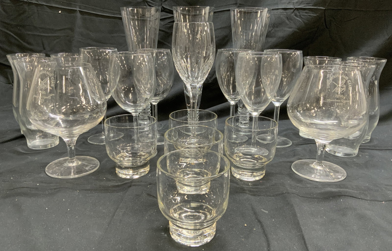 Lot 23 Glassware Vessels Barware: Lot Includes 6 wine glasses, 6 water glasses, 5 votives/candle holders, 3 champagne flutes, 2 monogrammed brandy sniffers (THE APAWAMIS CLUB 1992) & 1 Crystal wine glass. Champagne flutes measure appr
