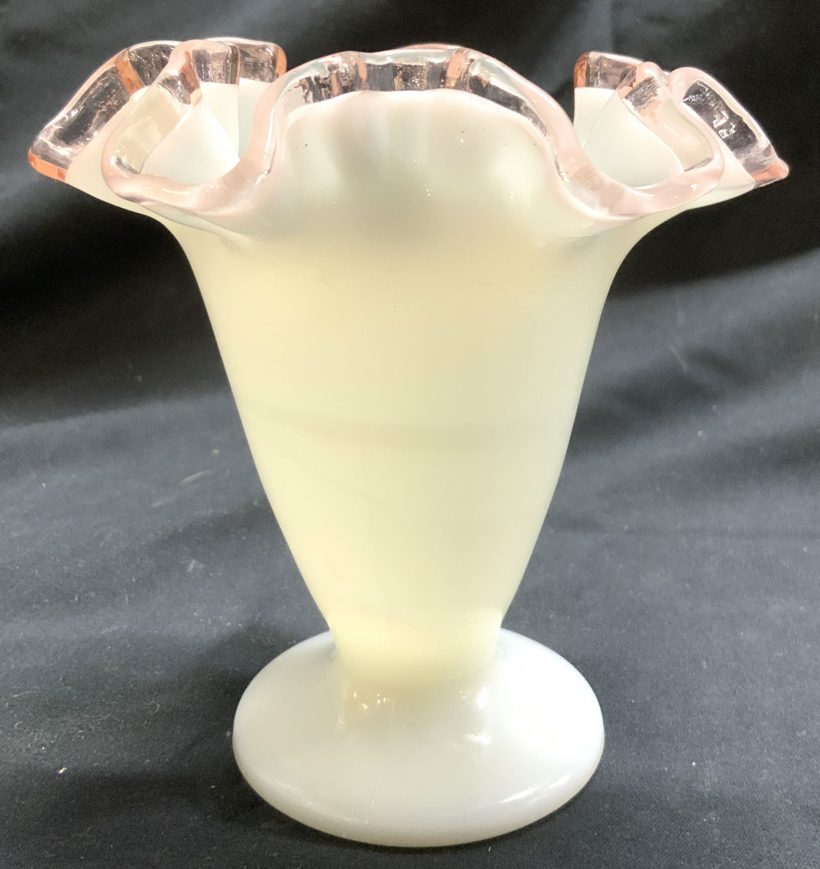 Milk Glass Handkerchief Glass Footed Vase: Vase is opaque white toned. Piece has pink toned rim. Vase is footed with circular form base. Piece has handkerchief style structure to rim. Vessel measures approximately 4.25 inches tall and wide. Va