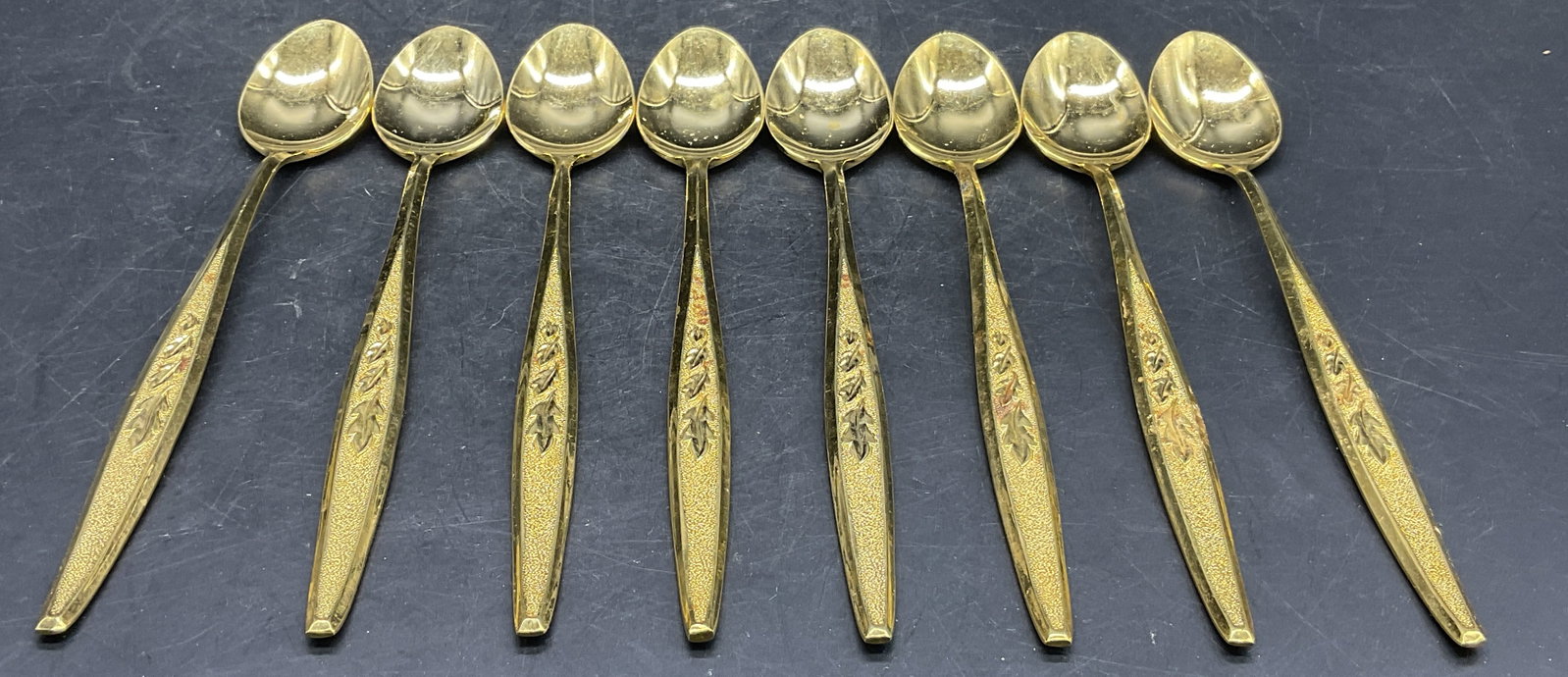 Set 8 REGENT STAINLESS Gold Tone Metal Spoons: Set 8 REGENT STAINLESS Gold Tone Metal Spoons. Undersides signed REGENT, STAINLESS, JAPAN. Measures approx. 7L x 1W in. Foliage motif to the handle. Discoloration and surface wear present. Tableware, 