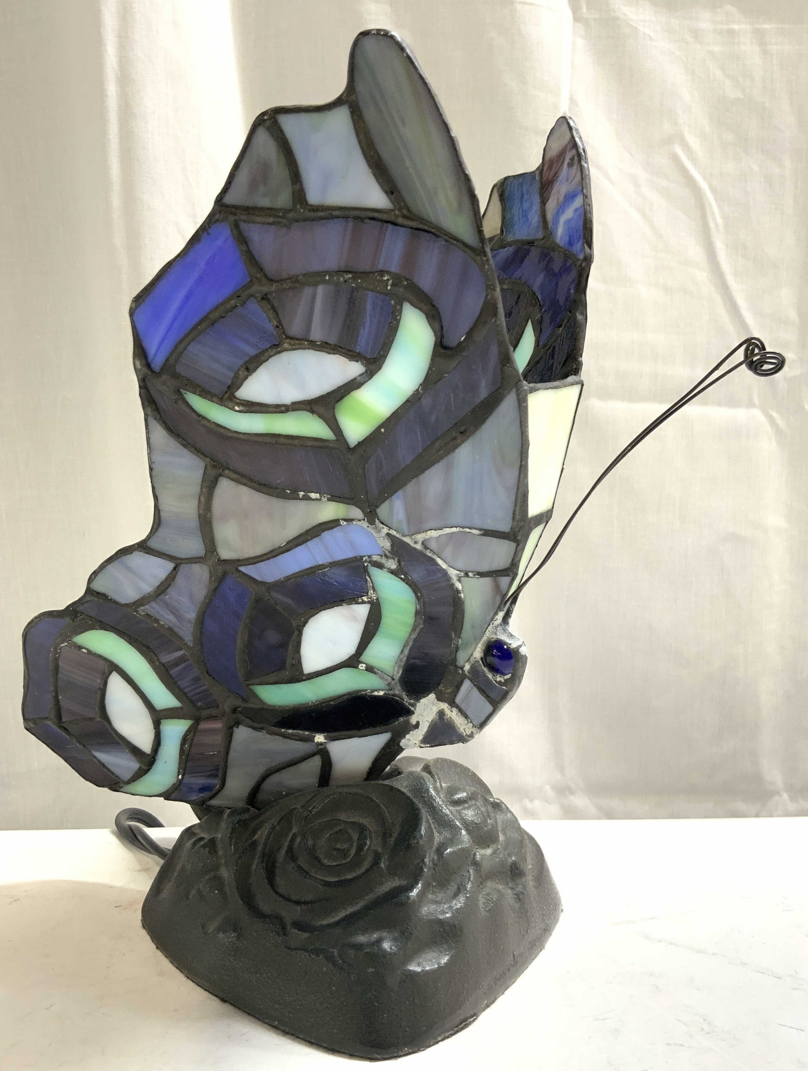 Tiffany Style Stained Glass Butterfly Lamp: Lamp is Tiffany Style. Lamp is in the form of a butterfly. Piece has stained glass detailed wings. Butterfly figure has antenna structures. Camp has dark brown toned floral detailed metal base. Lamp m
