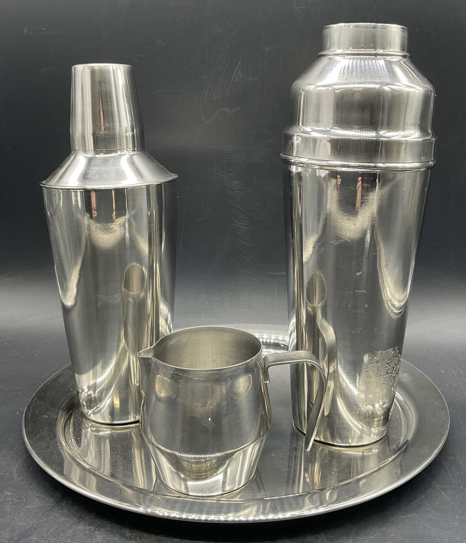4 Barware Silver Tn Metal Shakers, Pourer, & Tray: Four pieces of barware. Two shakers, one pourer, and one circular tray. Pieces are crafted from silver tone metal. Pourer underside reads MADE IN HONG KONG, STAINLESS STEEL. Tallest shaker measures ap