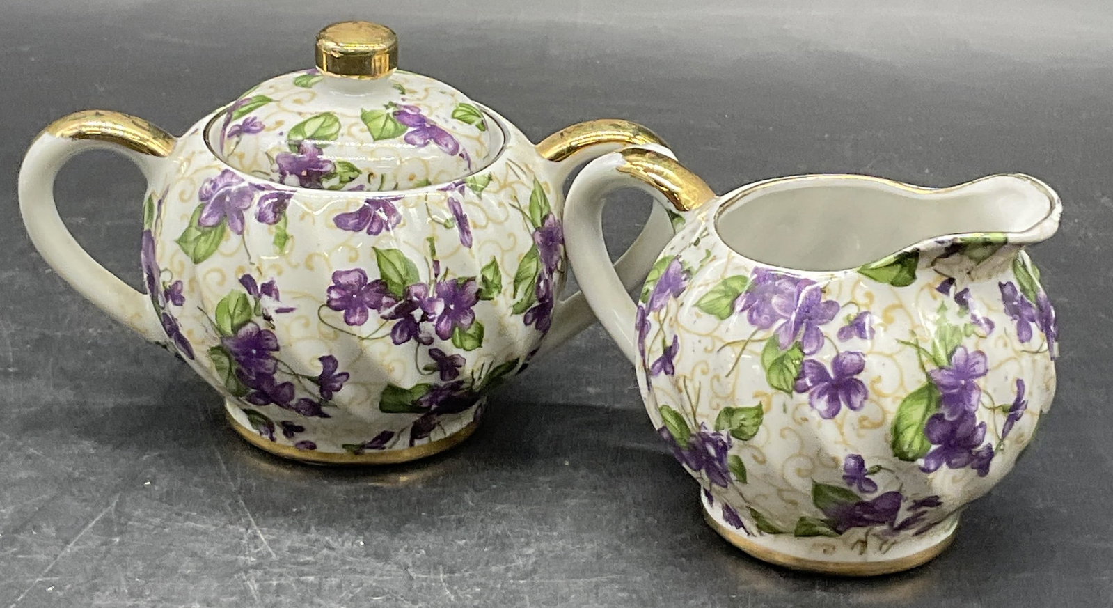 Set Purple Floral Porcelain Sugar Bowl & Creamer: Set consisting of a Purple Floral Porcelain Lidded Sugar Bowl & Creamer. White porcelain with deep yellow, purple & green florals and foliage to the design. Gilt line work to the pieces. Creamer measu