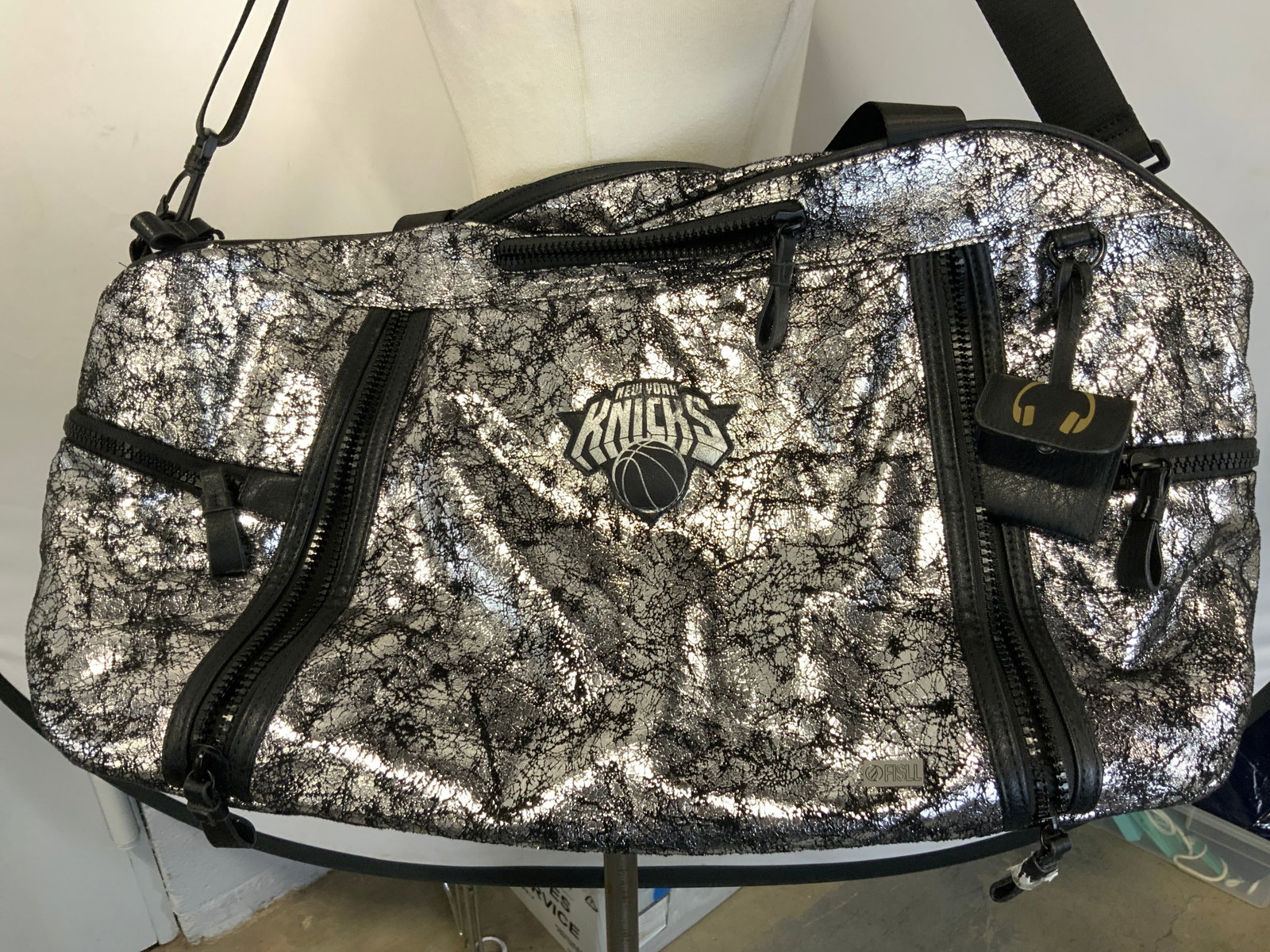 NEW YORK KNICKS Foil Gym Bag: Gym bag has NEW YORK KNICKS logo. Piece is black & silver toned with crackled foil detail. Piece has 5 exterior zippers, 2 interior zipper pockets, & 2 interior open pockets. Bag measures approximatel