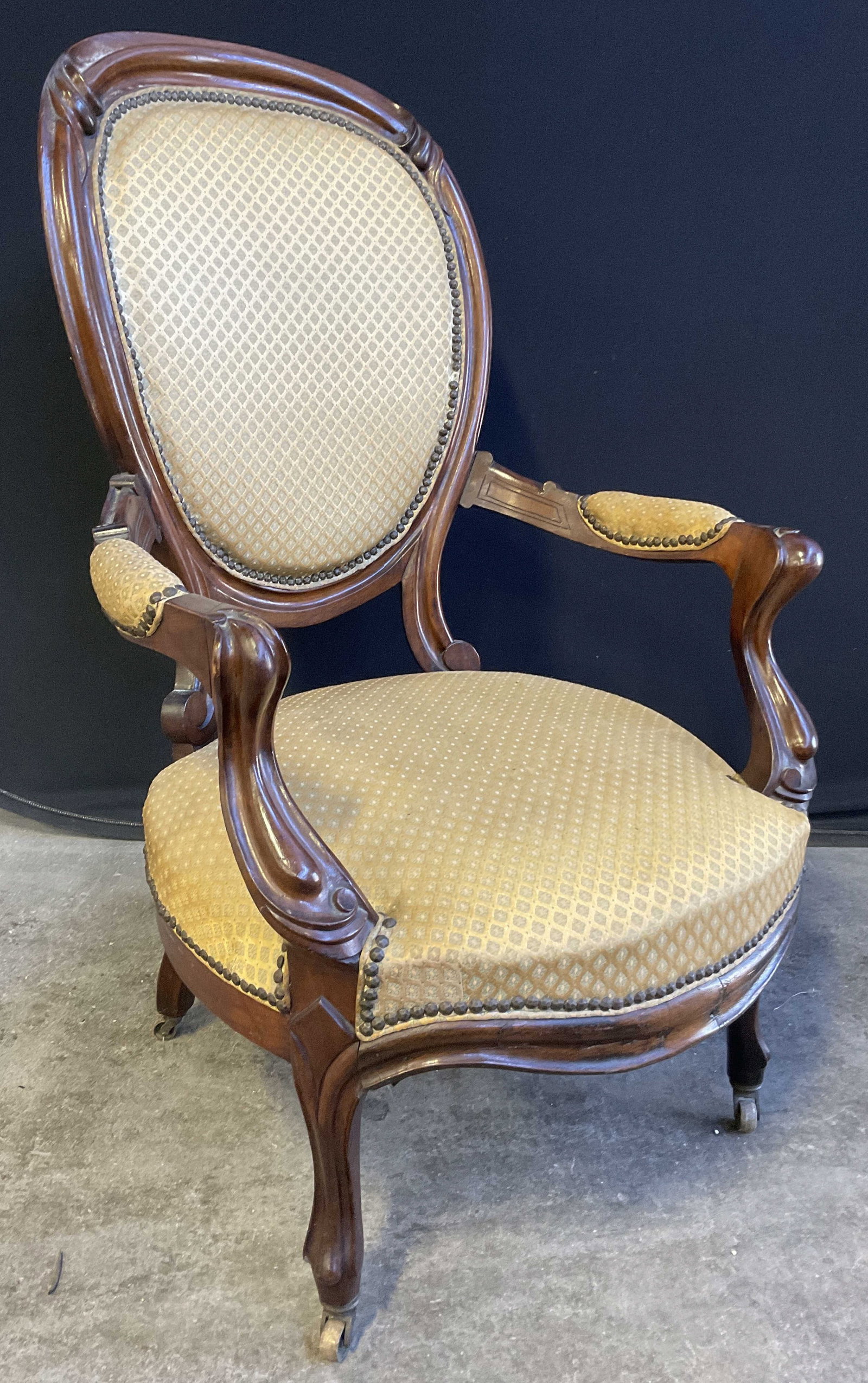 Antique Upholstered Parlor Chair On Casters: Antique, possibly vintage, upholstered parlor chair on casters. Carved wood frame. Metal nailhead details. Measures approx. 43H x 24.5W x 24D in. Discoloration, surface wear, losses, and separation pr