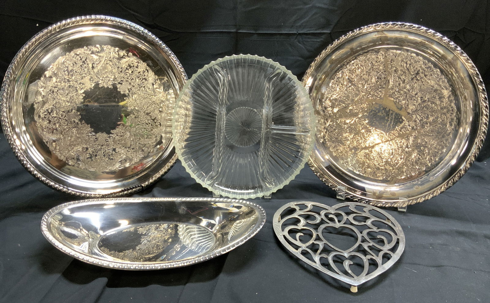 Lot 5 Silver Plate & Glass Tabletop: Lot includes 2 circular form silver plate tray with ornate scroll and leaf relief detail, 1 silver plate tray with oval form, 1 sectioned fluted glass tray and 1 heart form metal trivet. Circular tray