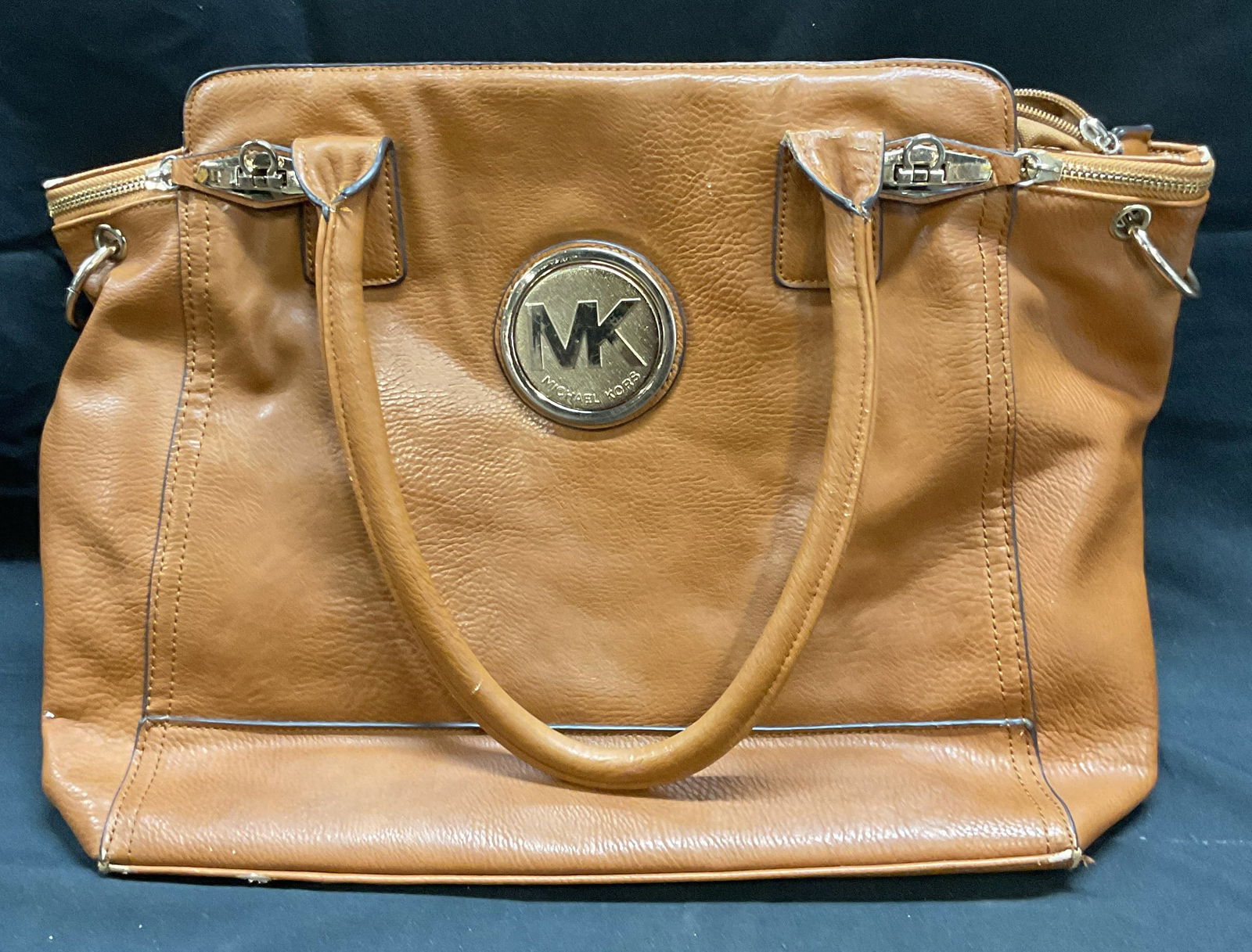 Tan Faux Leather Handbag: Tan faux leather handbag. Gold tone hardware. Multiple interior pockets. Label reads Michael Kors. Measures approx 13.5 x 5 x 12 inches. Wear to materials including scratching, peeling, and blemishes