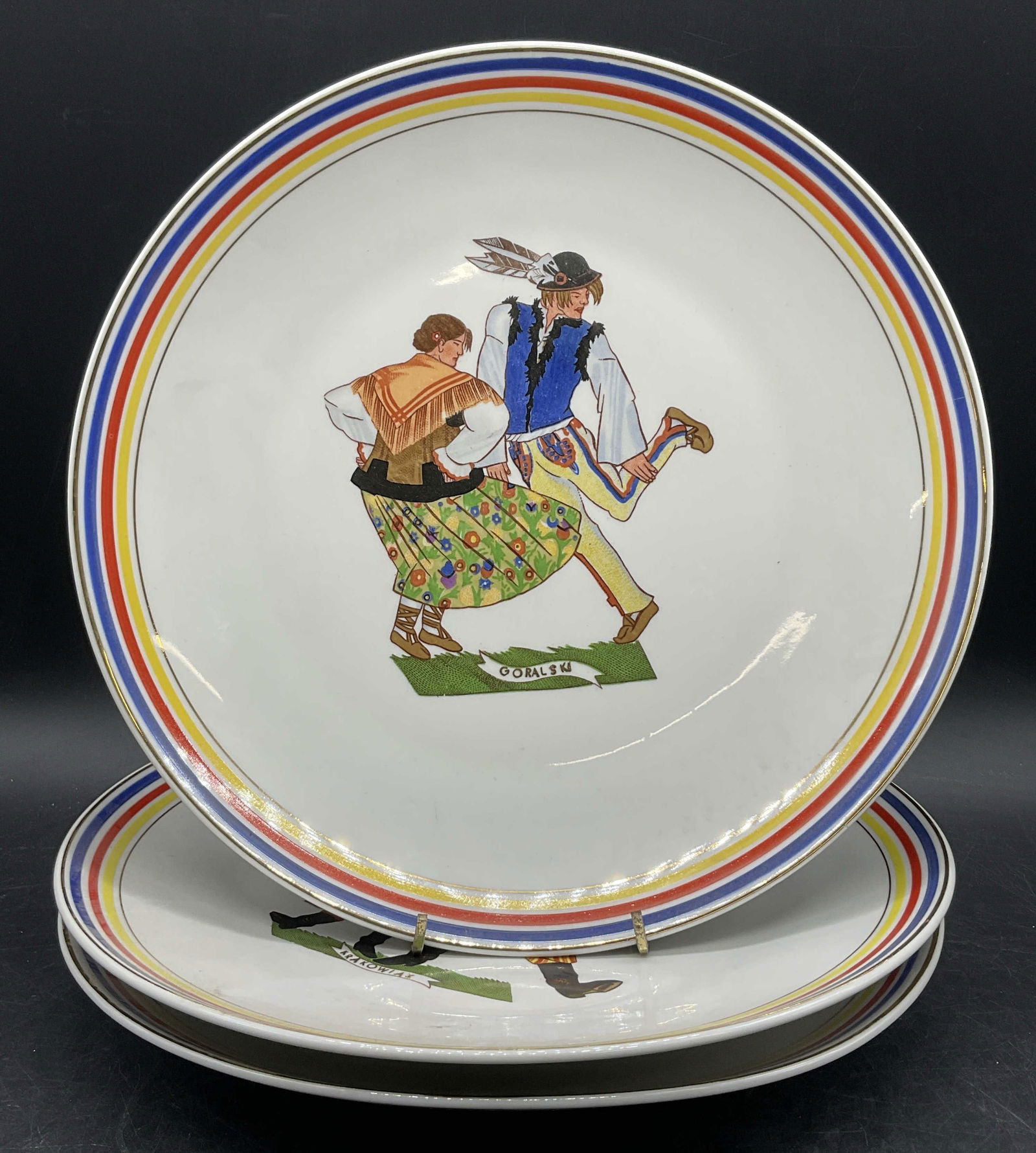 Lot 3 CMIELOW POLAND Vintage Folk Dance Plates: Three CMIELOW POLAND Vintage Folk Dance Motif Plates. Plates feature people in multicolored clothing to the center engaged in folk dances. Gilt rims. Measures approx. 11.5L x 11.5W in. Drilled holes t