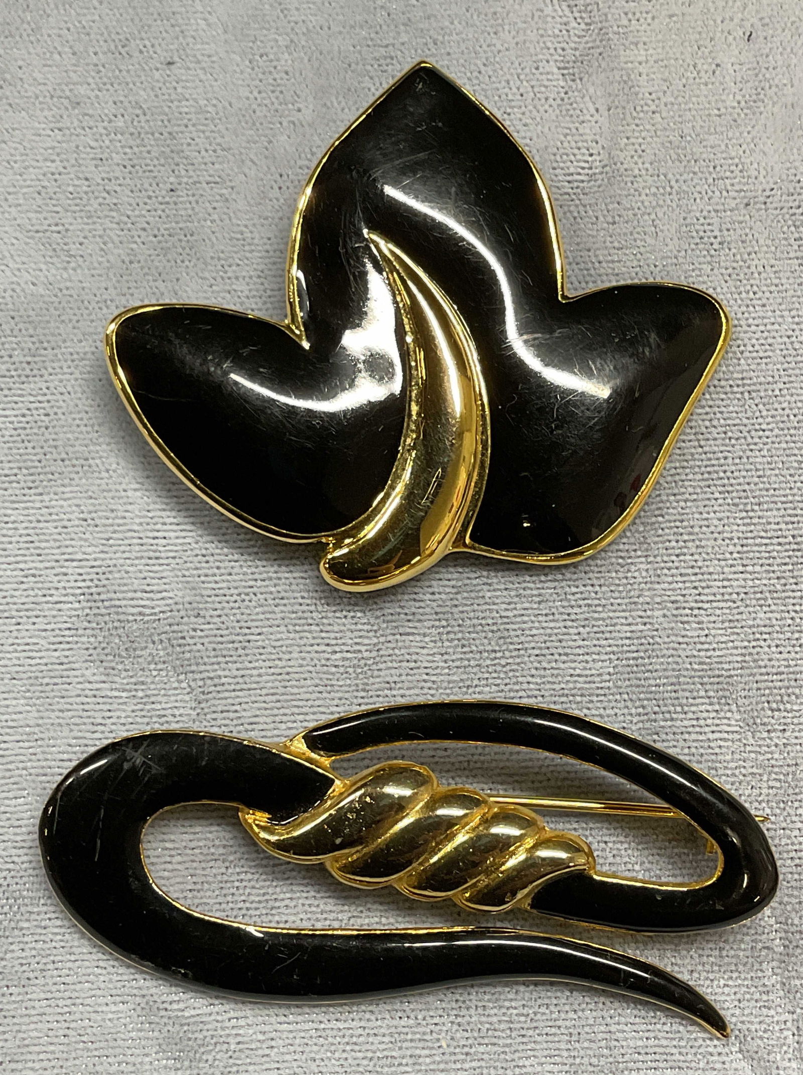 Lot 2 Vintage Gold Tone Black Enamel Brooches: Lot of 2 vintage gold tone and black enameled brooch. Flower or leaf motif and squiggle motif. Hinge pins on undersides. Larger brooch measures approx 2.5 inches long. Some wear to each including scra