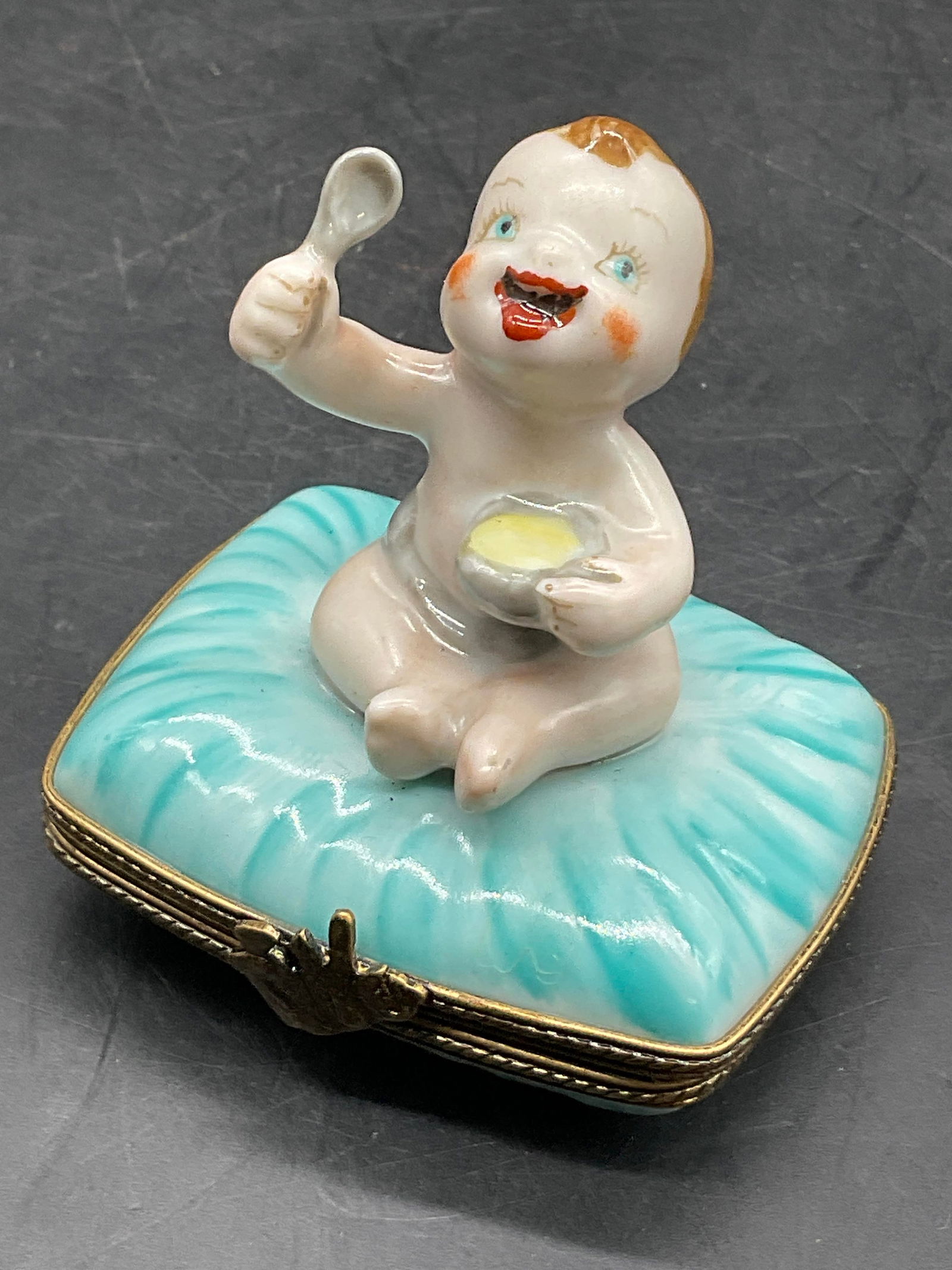 LIMOGES FRANCE Hand Painted Porc Baby Trinket Box: LIMOGES FRANCE Hand Painted Porcelain Baby Motif Trinket Box. Subject is a baby sitting atop a pillow eating from a bowl. Opens with metal hinge hardware. Underside signed PEINT MAIN LIMOGES FRANCE. S