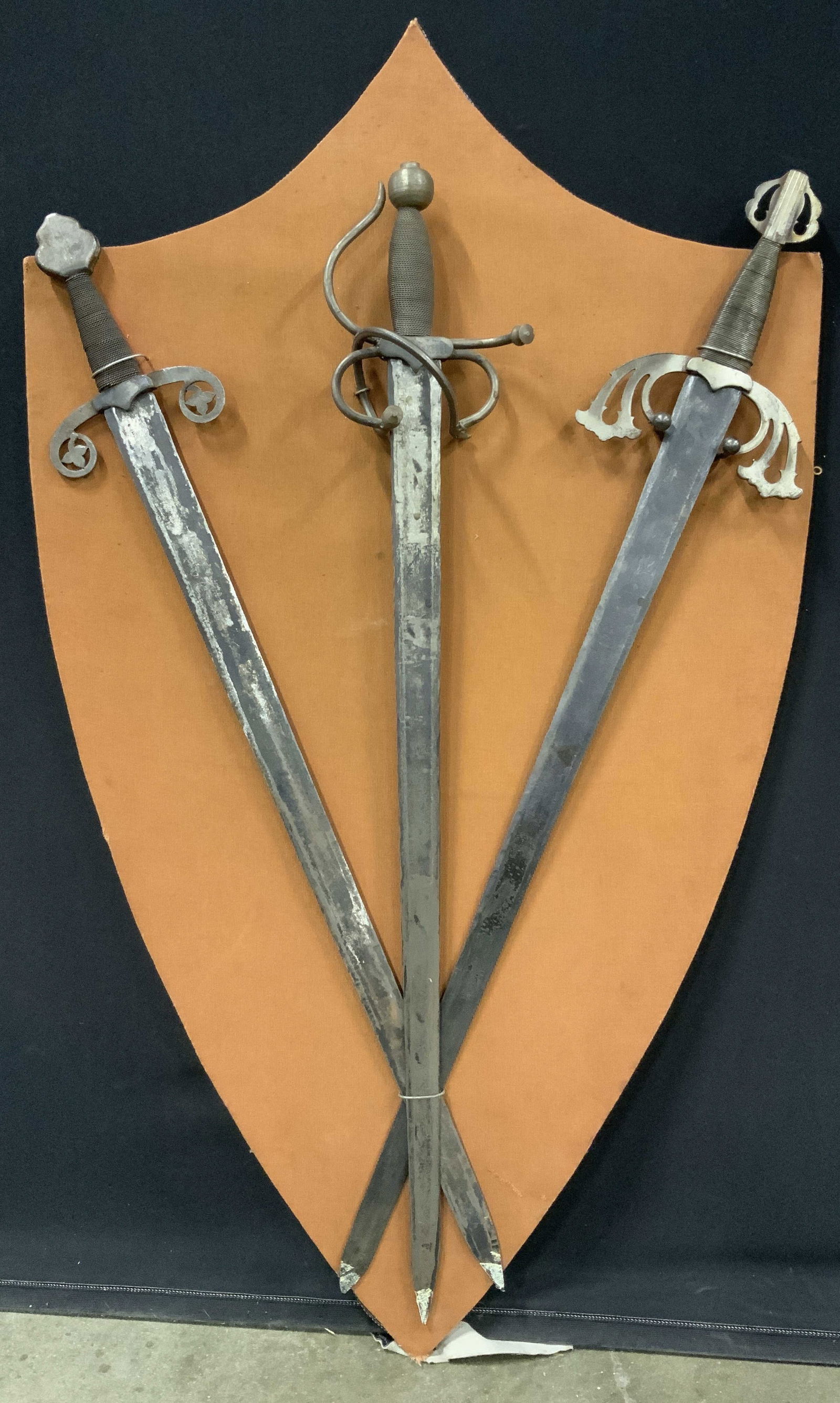 3 Mounted Cantar de Mio Cid Metal Replica Swords: 3 mounted swords on shield shaped wooden back board. These three swords are replicas from the Mexican epic poem Cantar de Mio Cid. Two of the swords have names , Tizona and Colada. Back board has an e