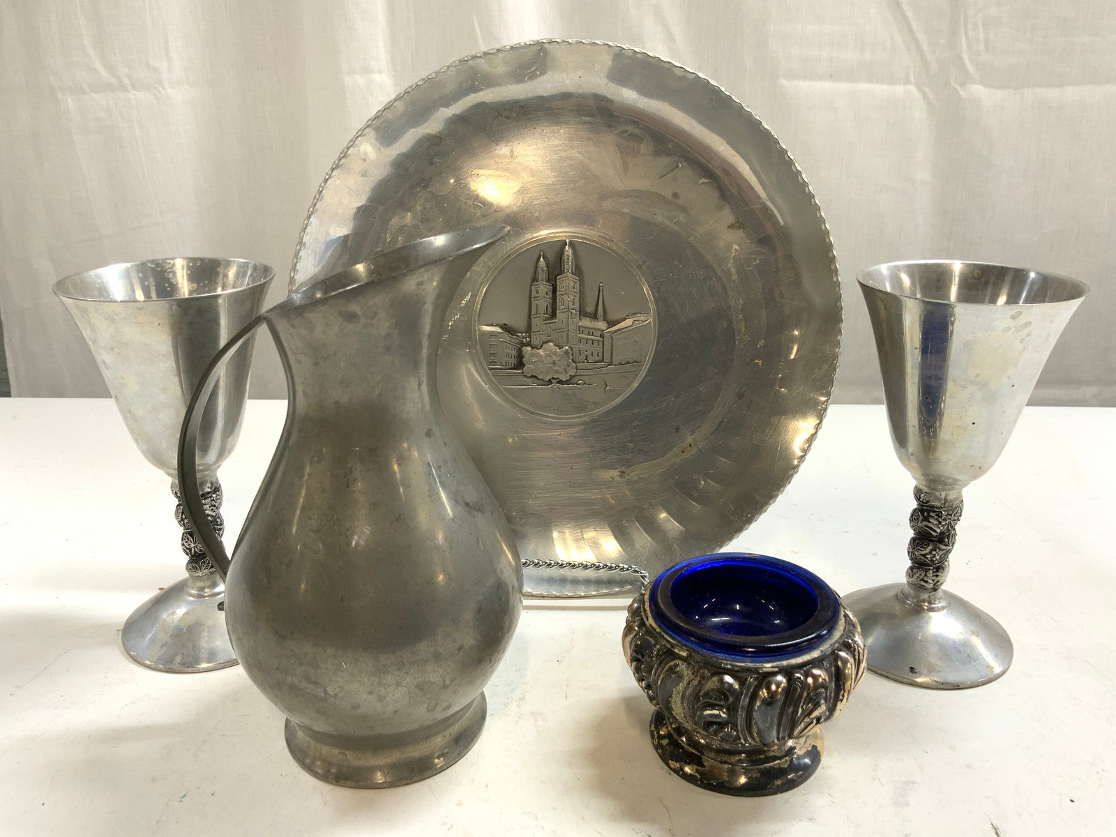 5 Pewter Accs, Signed Zurich, Raimond Italy & More: Lot includes 1 plate reads ZURICH GROSSMUNSTER, 2 Chalices trademarked RAIMOND ITALY, 1 Vessel with cobalt blue glass insert, & 1 pitcher inscribed JEKA TIEL HOLLAND. Plate measures approximately 8.25