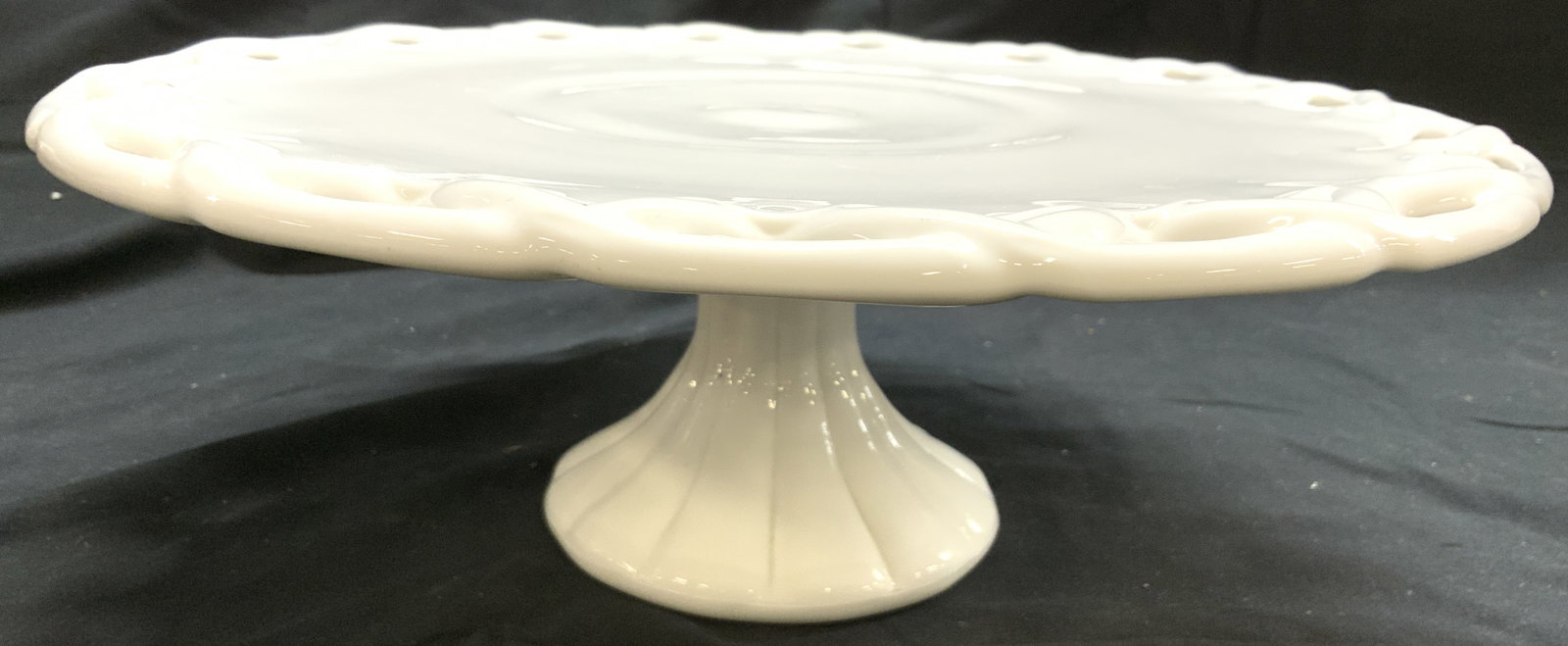 Art Glass Pedestaled Cake Stand Milk Glass: Cake stand is white toned and glass, milk glass. Piece has pierced detail at edges. Pedestal is scalloped. Cake tray has circular form. Piece measures approximately 14 inches diameter 5 inches tall. C