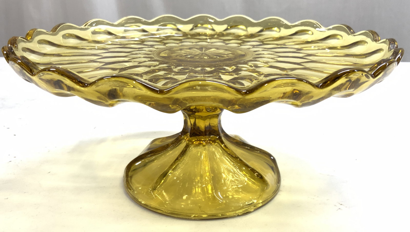 Amber Glass Pedestaled Cake Stand: Cake stand is amber glass. Piece is pedestaled. Cake stand has Geometric relief detail. Edges of rim have rounded teeth. Base has circular form. Piece measures approximately 10 inches wide, 4.5 inches