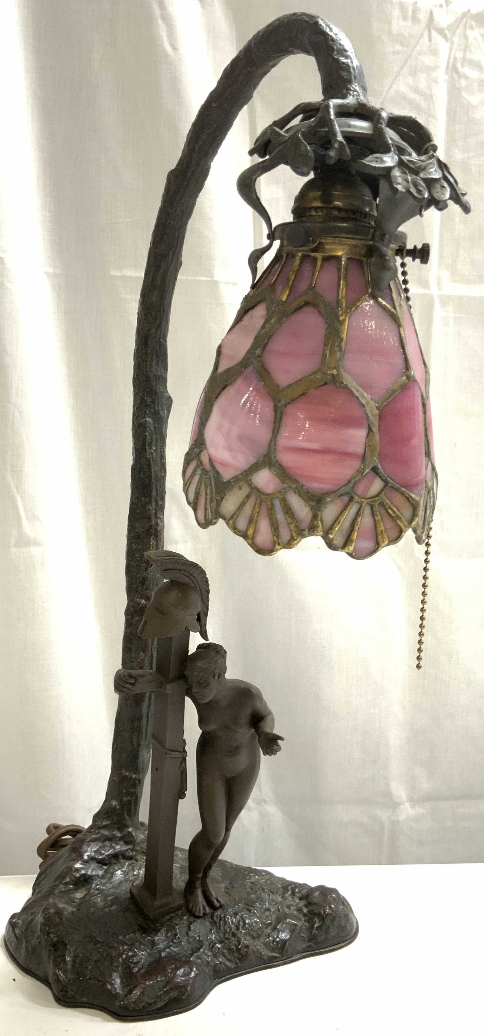 Bronze Female Nude Tiffany Style Table Lamp: Lamp base has female nude figure standing next to post with Helmet on top. Lamp base appears to be bronze. Piece has pink toned Swag glass shade. Lamp also has beaded metal pull switch. Piece measures