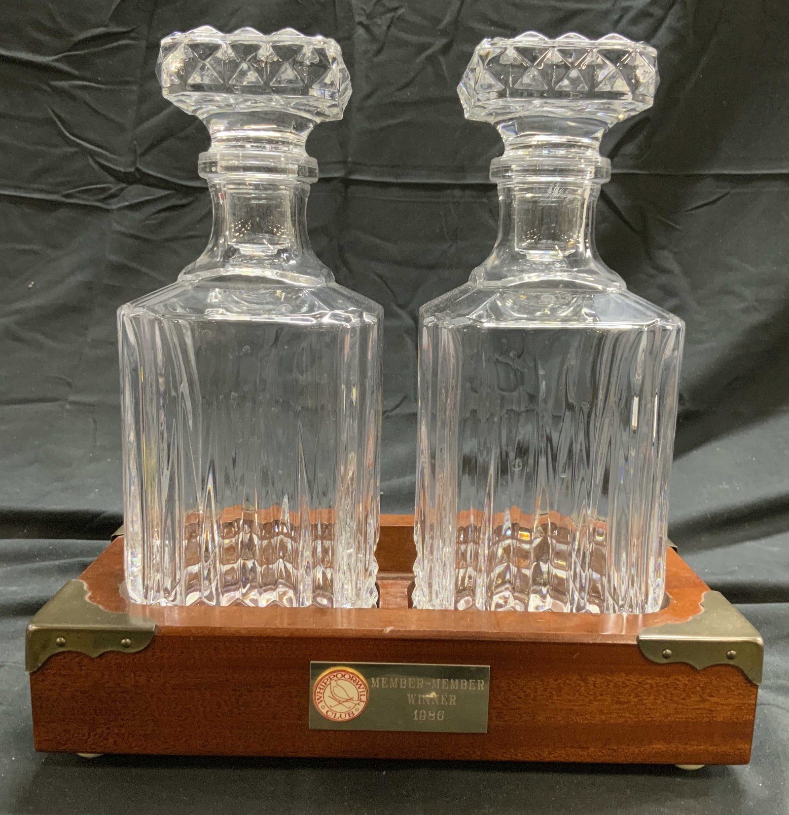 Pr Gls Decanters & Whippoorwill Club Wood Holder: Plaque on front reads Whippoorwill Club Member Member Winner 1986. Tag on underside reads PRIZE POSSESSIONS. Lot includes 1 wooden decanter holder with metal overlay to corners & 2 Art glass decanter 