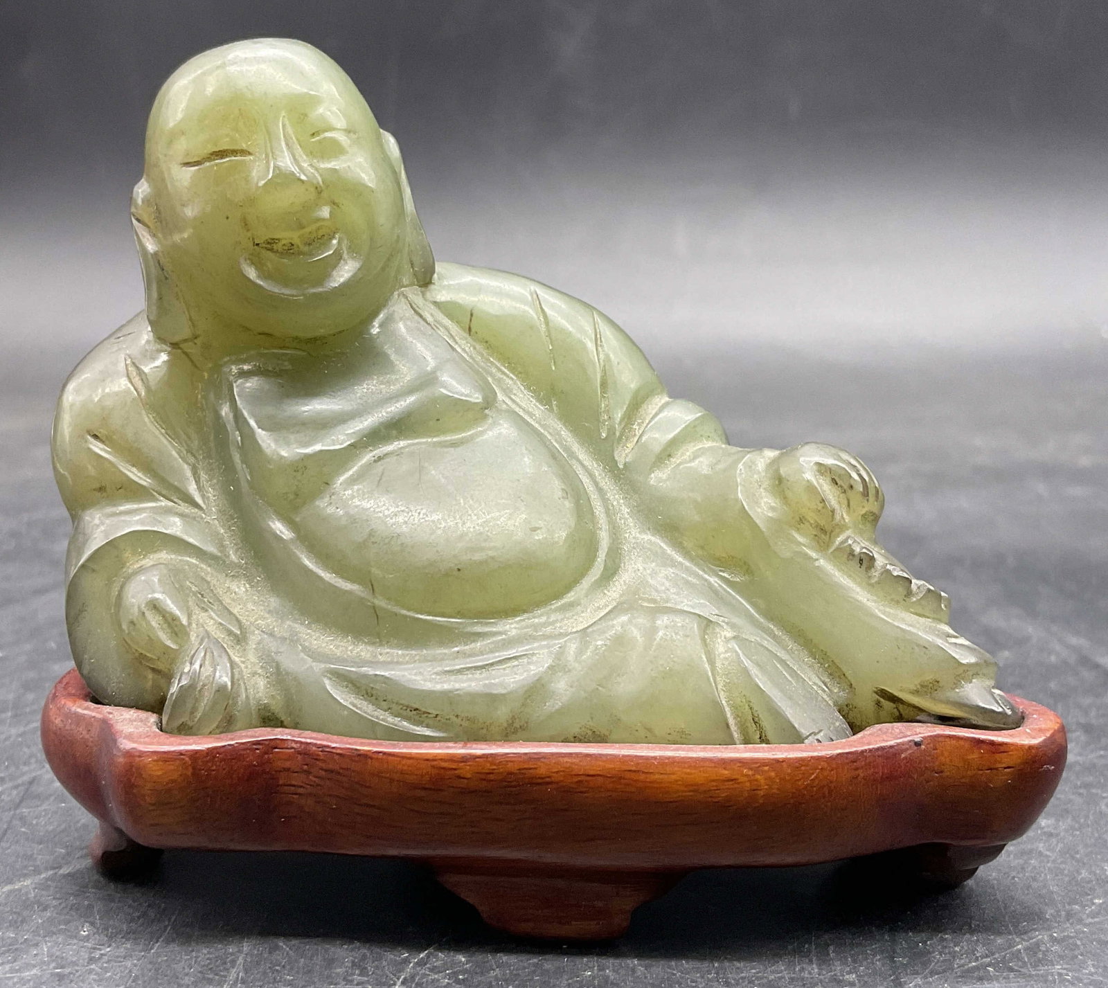 Green Stone Laughing Buddha Figural W/ Stand: Green Stone Laughing Buddha Figural W/ Wooden Stand. Sticker to underside of Buddha reads Made in Peoples Republic of China. Together pieces measure approx. 3H x 3.5W in. Residue, chipping, and surfac