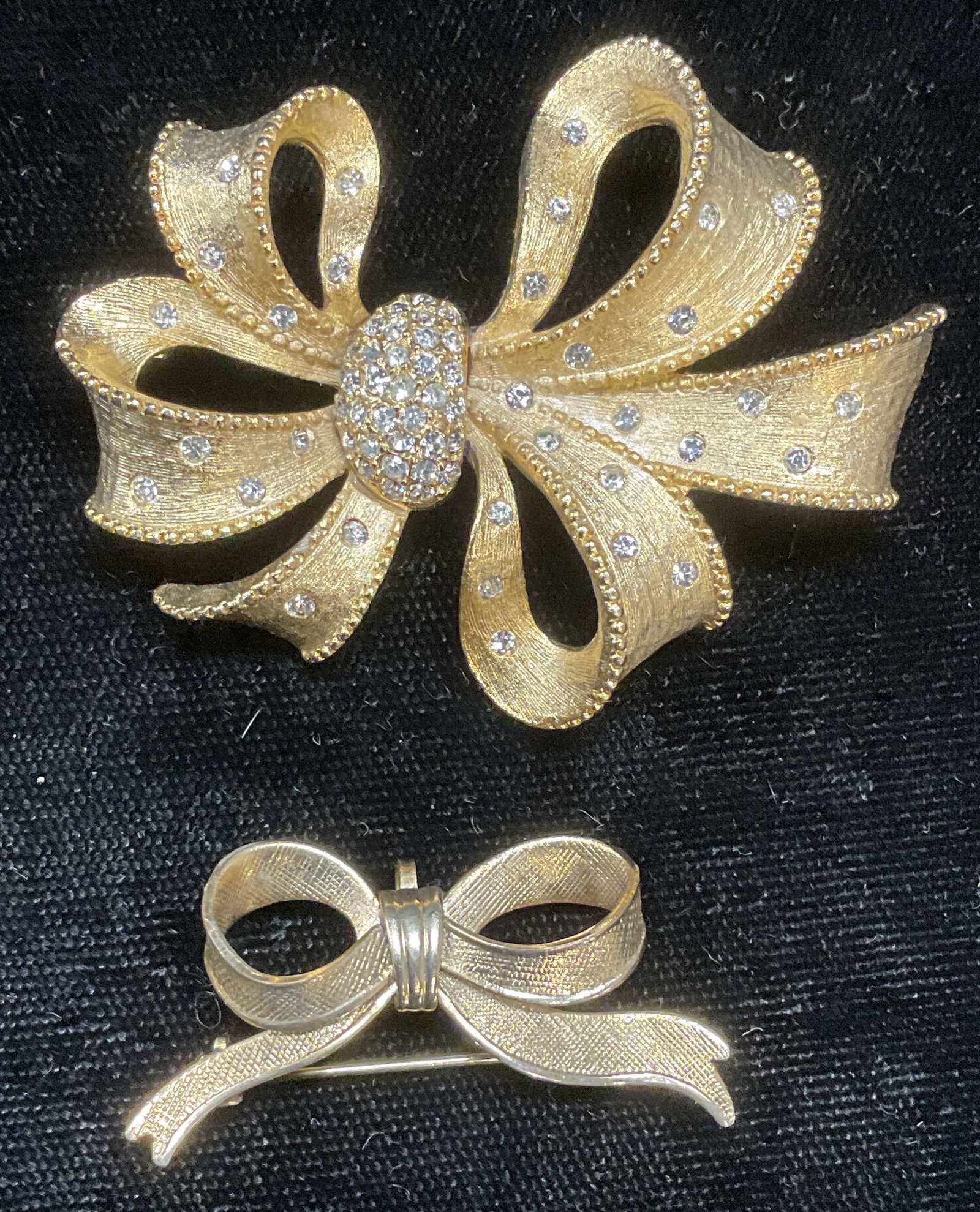 Lot 2 Vintage Gold Tone Ribbon Bow Brooches: Lot of 2 vintage gold tone ribbon bow motif brooches. Textured and crystal details. Hinge pins on undersides. Larger piece measures approx 1.75 x 2 inches. Some wear to each including scratching obser