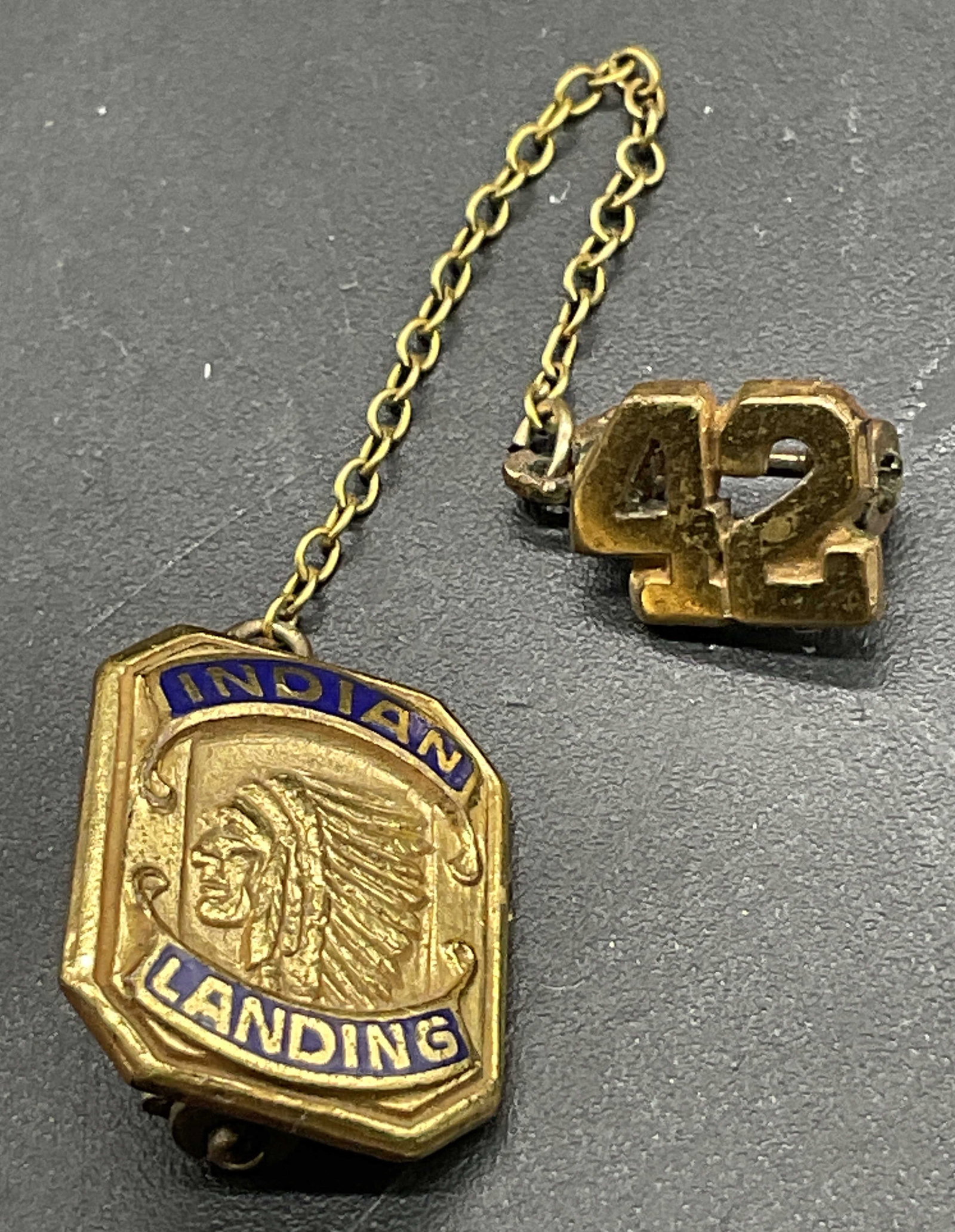 INDIAN LANDING 42 Monogram HVM Pin: INDIAN LANDING 42 Monogram HVM Pin. Features the number 42 and a Native American head that reads INDIAN LANDING. Crafted from gold tone metal with blue enamelwork design. 2.5L x .5W in. Metal pin clos