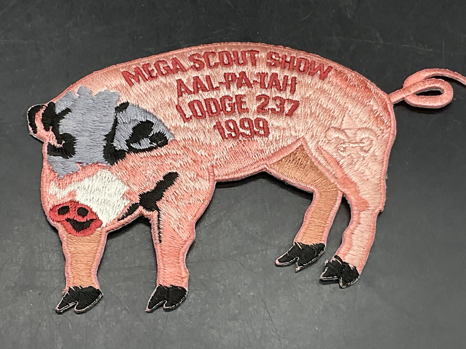 MEGA SCOUT SHOW AALPATAH LODGE 297 1999 Pig Patch: MEGA SCOUT SHOW AAL PA TAH LODGE 297 1999 Fabric Pig Patch. Primarily pink, gray, and black hues. Measures approx. 5.5W x 3.5L in. Surface wear consistent with age, use, and exposure. Animal patch, fa