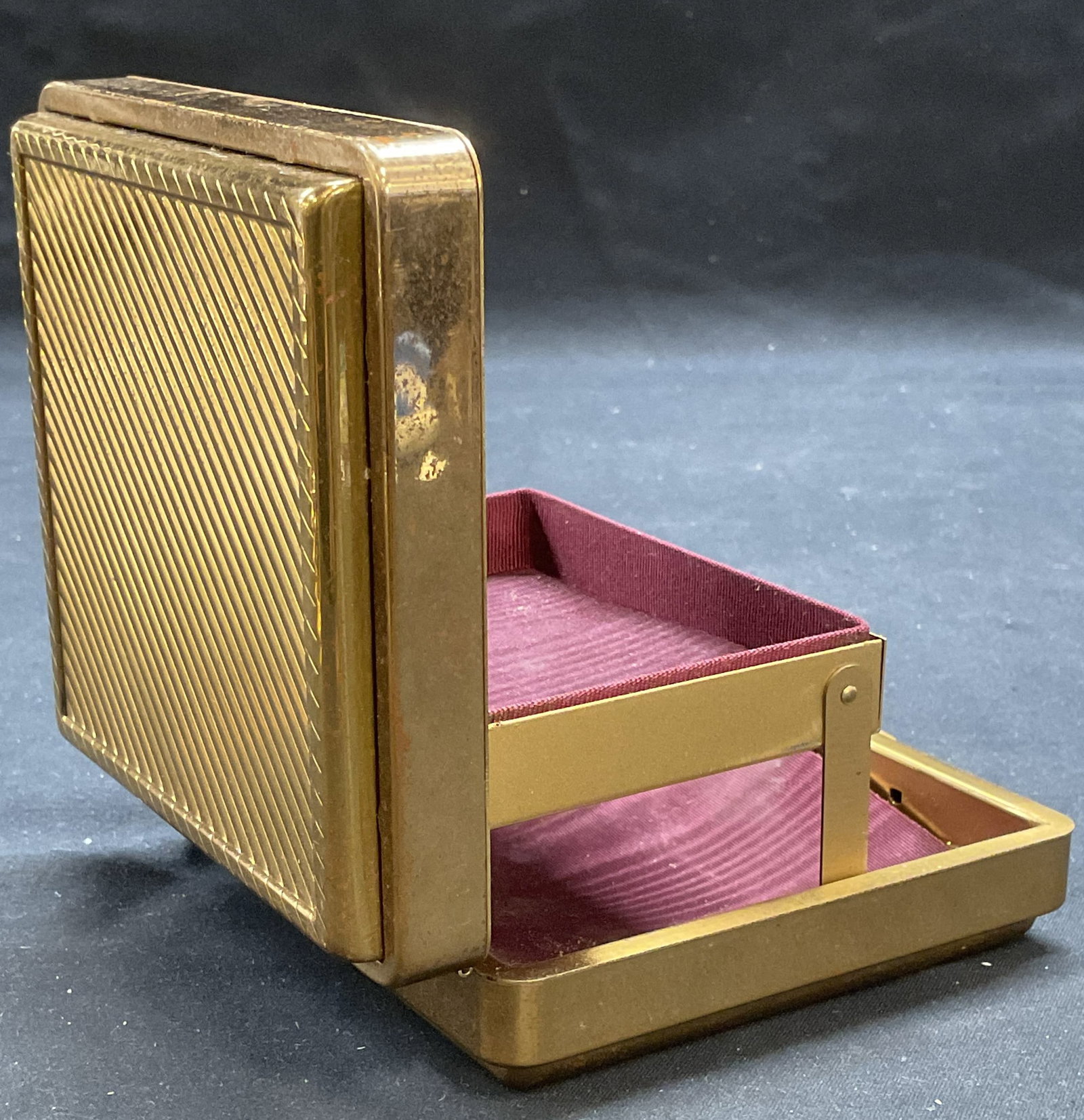 Vintage Gold Tone Folding Tray Jewelry Box: Vintage gold tone jewelry box. Ribbed stripes outer case. Interior with tray that folds out when opened. Purple fabric lining, not attached to case. Box measures approx 3.75 x 3.75 x 1.5 inches. Piece
