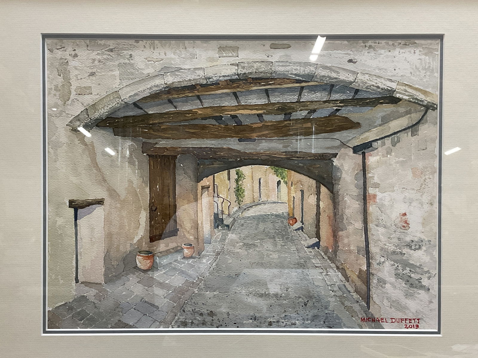 Michael Duffet Street Scn Watercolor Painting 2013: Framed, matted, and signed watercolor painting by Michael Duffet. Signed and dated 2013 at lower right. European street scene, roadway under bridge with door. Framed in silver leaf wood frame. Not exa