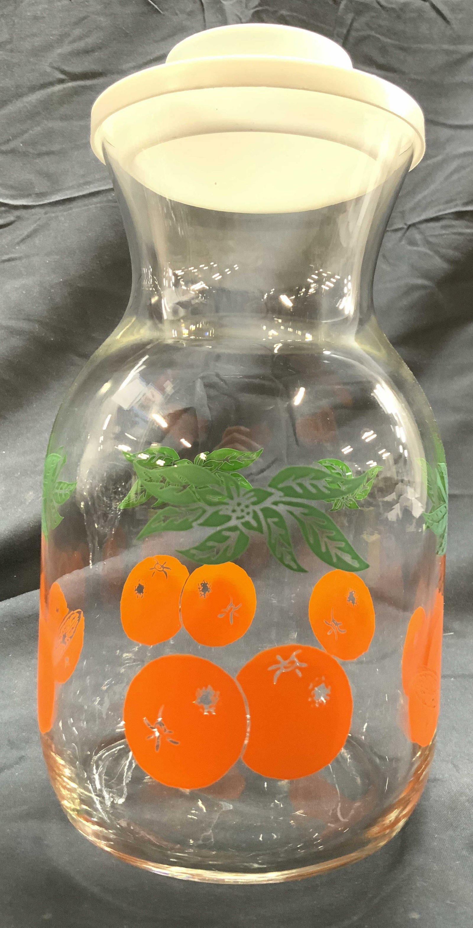 Art Glass Orange Juice Container w Lid: Orange juice container is glass. Piece has vase form with oranges and leaf detail. Vessel includes white toned lid. Piece measures approximately 8.75 inches tall 4.5 inches deep. Juice container, oran
