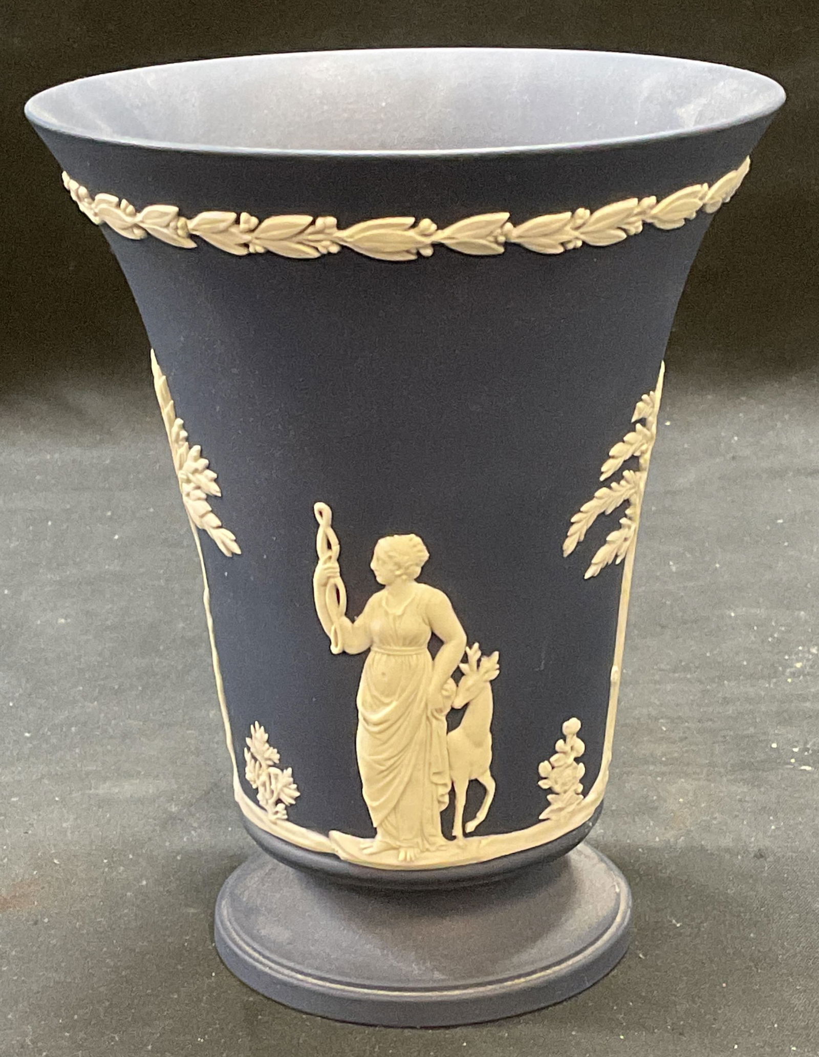 Wedgwood Dark Blue Jasperware Vase, England: Wedgwood Jasperware vase in dark blue. White bas relief Grecian figural motifs with botanical wreath rim. Signed on underside. Made in England. Measures approx 4.75 x 6 inches. Some wear including scr