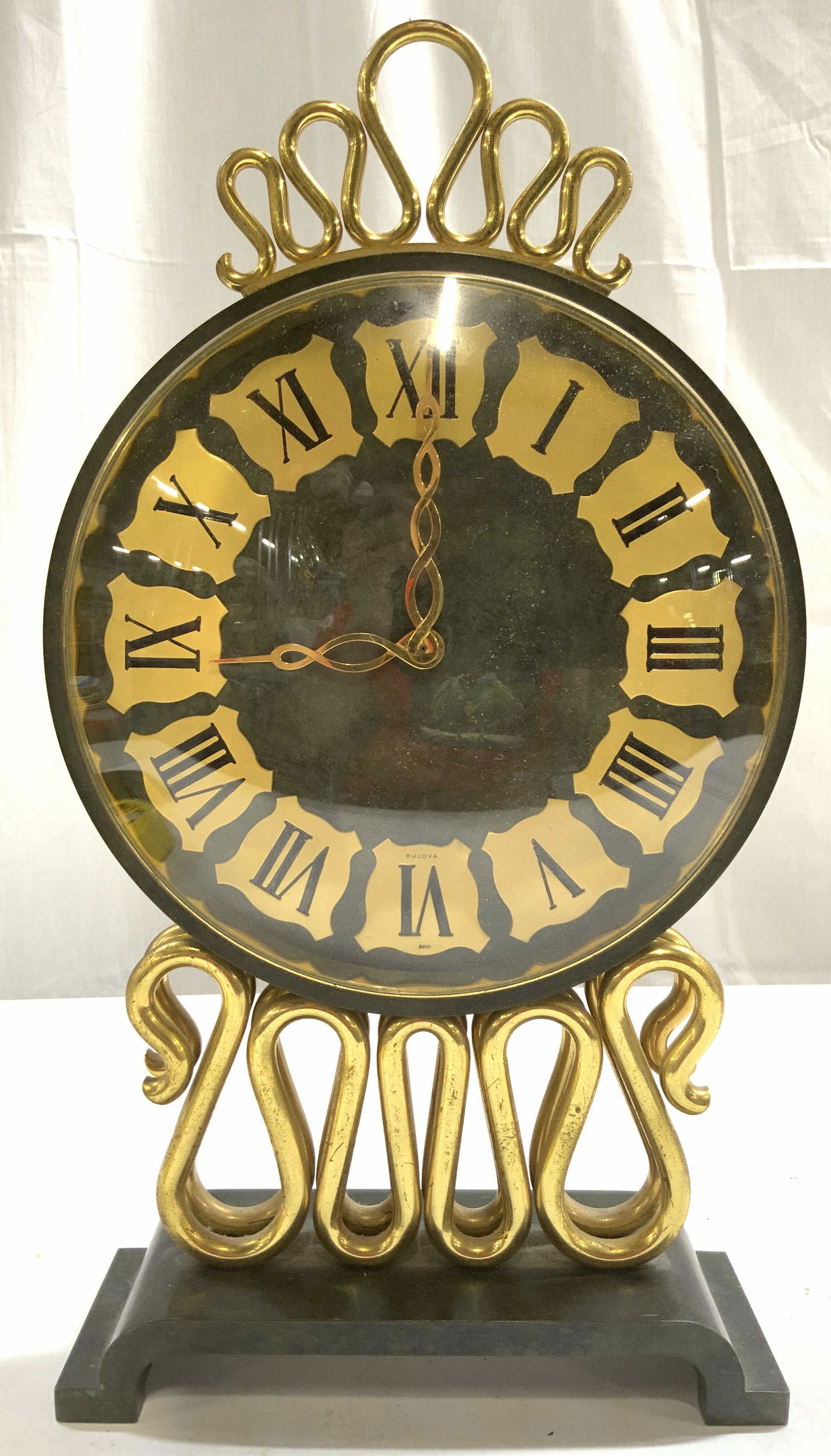 BULOVA of Switzerland Patinated Bronze Mantel Clock: Clock face reads BULOVA SWISS. BULOVA timepiece. Roman numeral face incased in patinated bronze with glass front supported by an undulating brass stylized base and matching crown. Clock face has Roman
