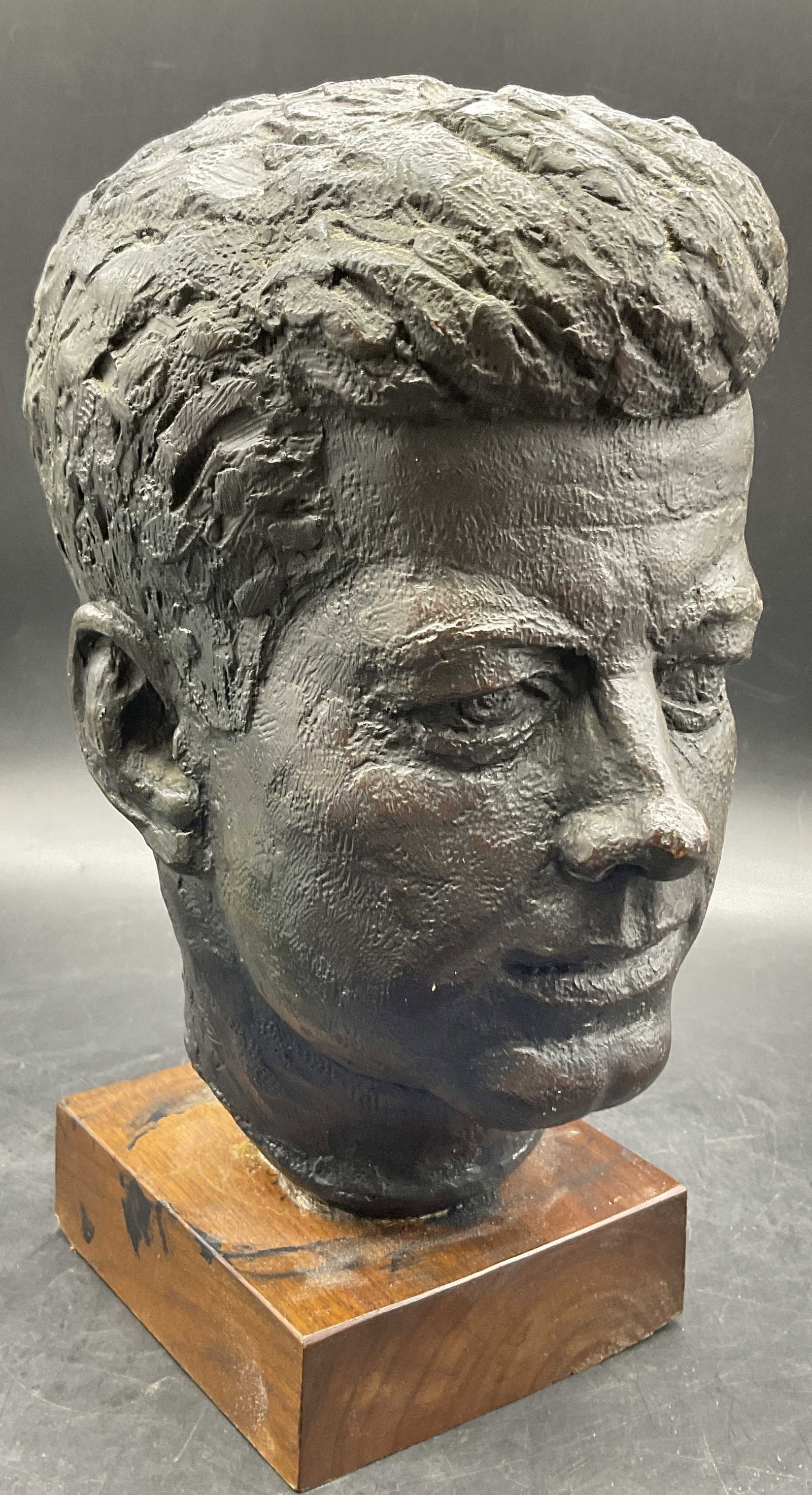 AUSTIN PRODUCTION 64 JFK Metal Bust on Wood: AUSTIN PRODUCTION 64 JFK Metal Bust on Wood. John F. Kennedy metal bust affixed to a wooden stand. Side is signed AUSTIN PRODUCTION 64. Measures approx. 10.5H x 5W in. Measured at widest point. Residu