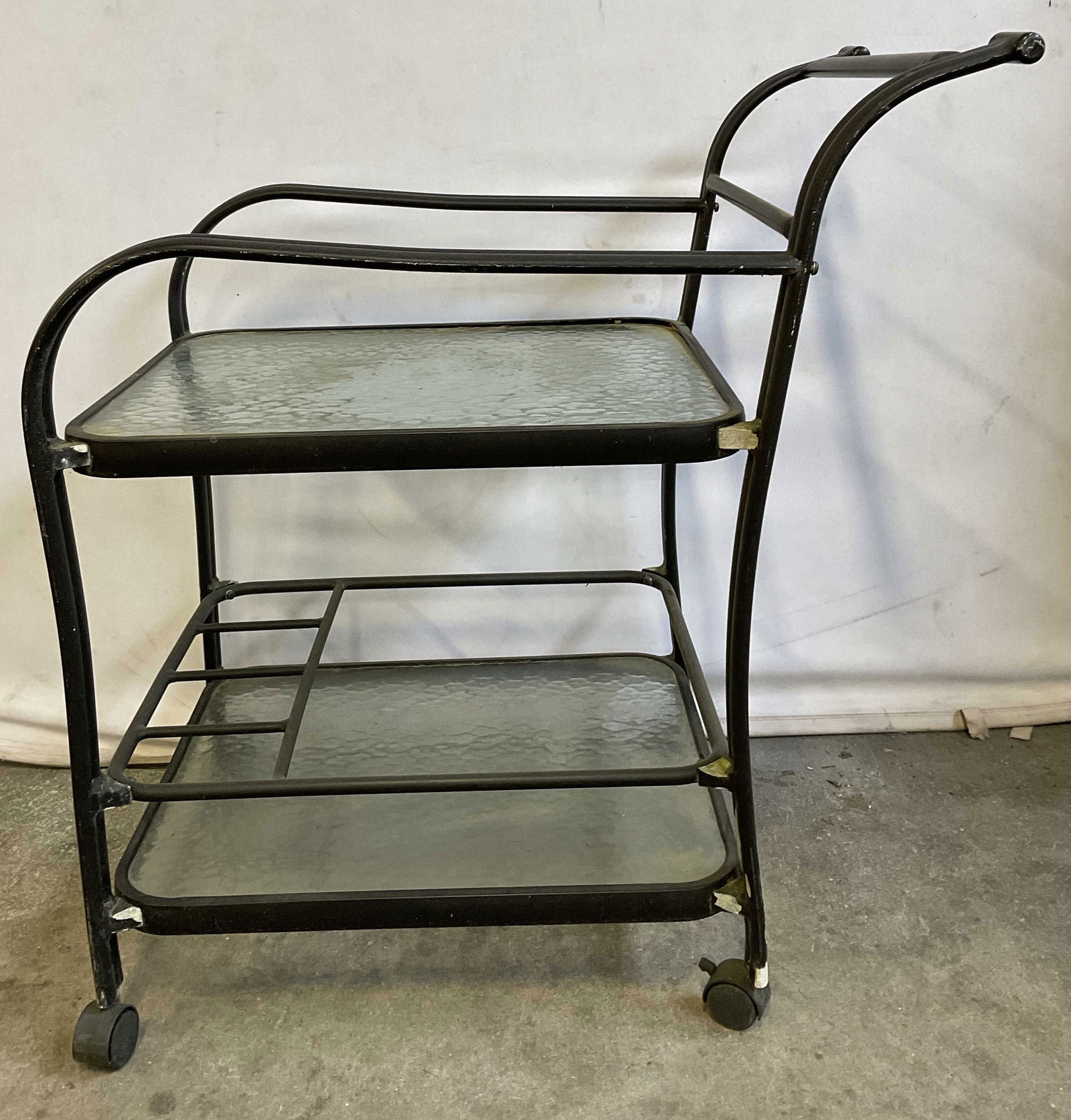 Vintage Black Metal Tiered Glass Bar Cart, Wheeled: Vintage Black Metal Tiered Glass Bar Cart on Wheels. Two tiered. Handle to one side. Measures approx. 36.5H x 22.75W x 32D in. Wheels need adjustment. Discoloration, denting, and losses to finish pres