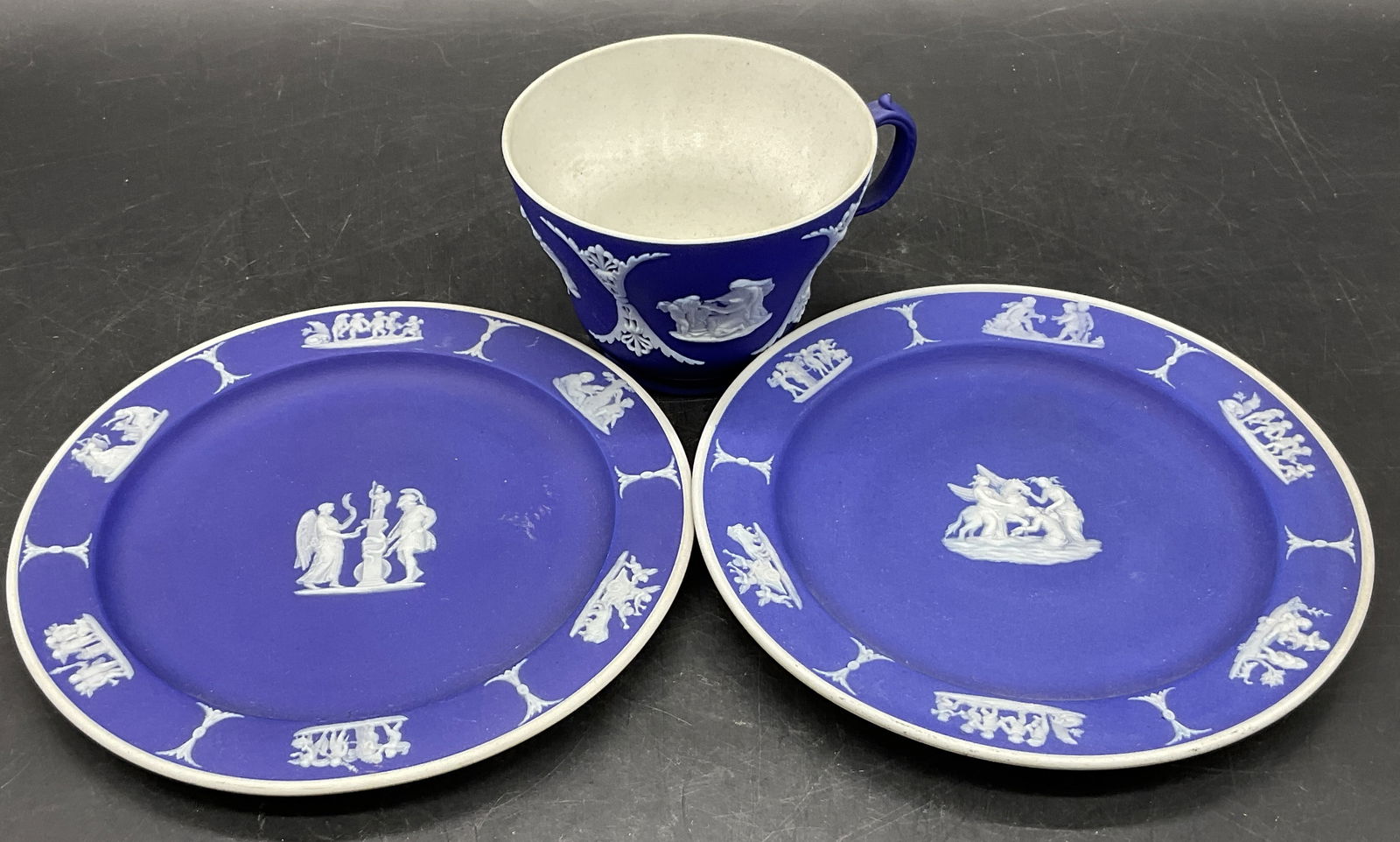 Lot 3 WEDGWOOD ENGLAND Blue Jasperware Cup+: Lot 3 WEDGWOOD ENGLAND Cobalt Blue Jasperware Plates & Cup. Pieces feature raised Greco Roman motifs. Plates measure approx. 6L x 6W in. Surface wear and discoloration present. Writing to undersides.