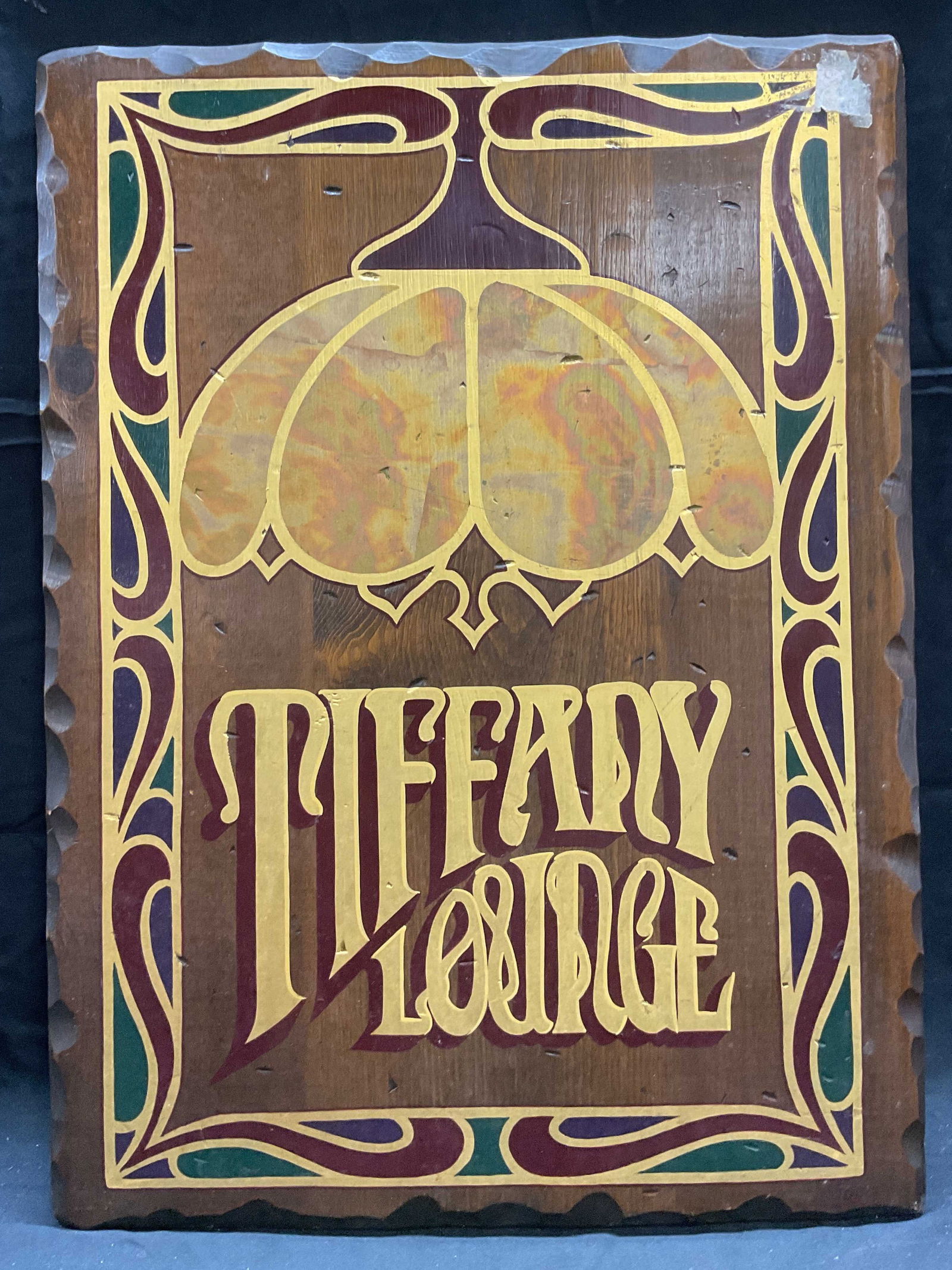 Tiffany Lounge Hand Painted Gilt Wood Sign: Tiffany Lounge wood sign. Hand painted with gilt decoration. Tiffany lamp motif with art nouveau style border in red, purple and green. Measures approx 24 x 17.5 inches. Some wear including scratching
