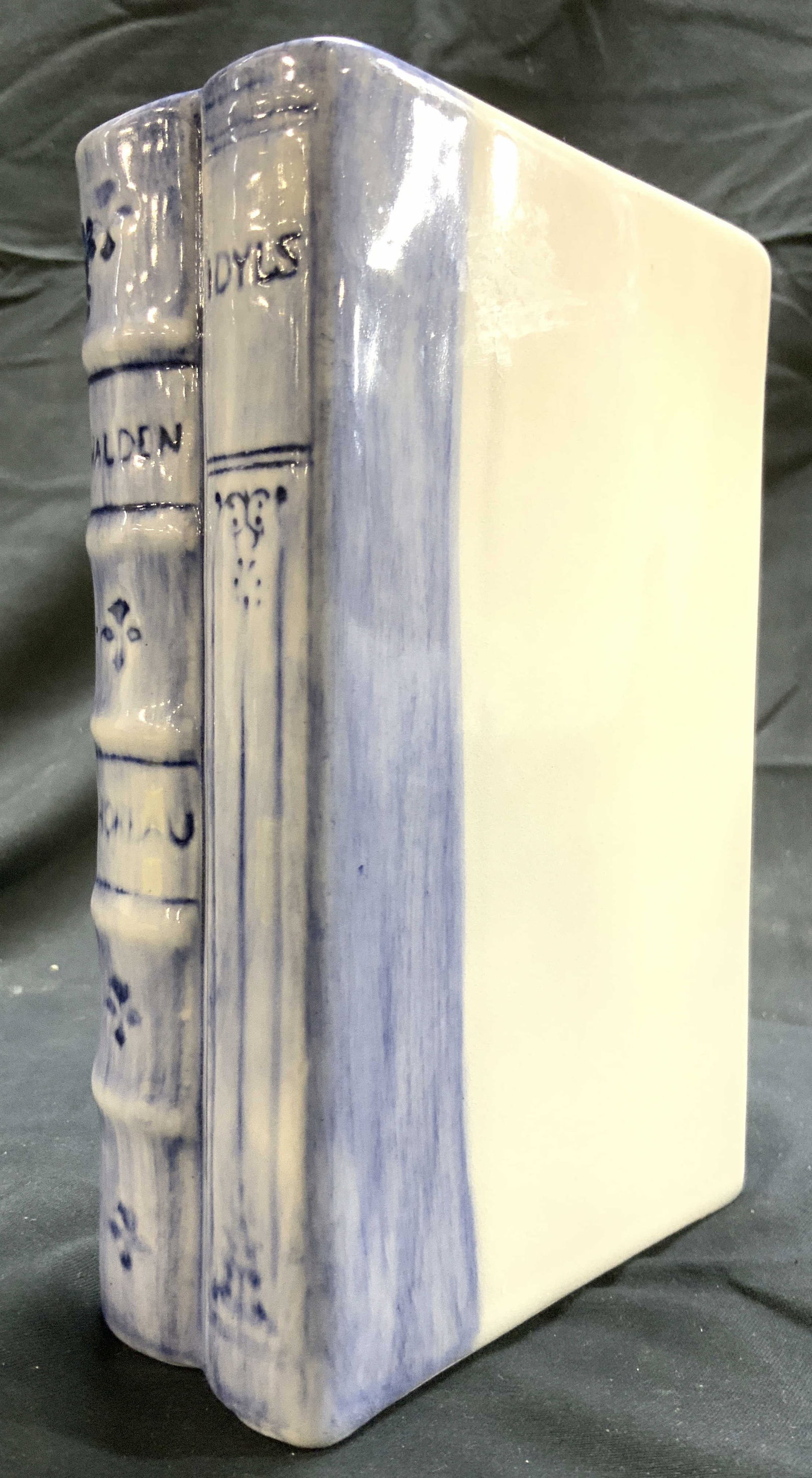 NS GUSTIN CO Vtg Porc Books Bookend: Tag on underside reads NS GUSTIN CO. Bookend is porcelain. Piece is white and light blue toned. Books figure reads WALDEN & IDYLS on spines. Piece has floral and leaf detail. Piece measures approximat