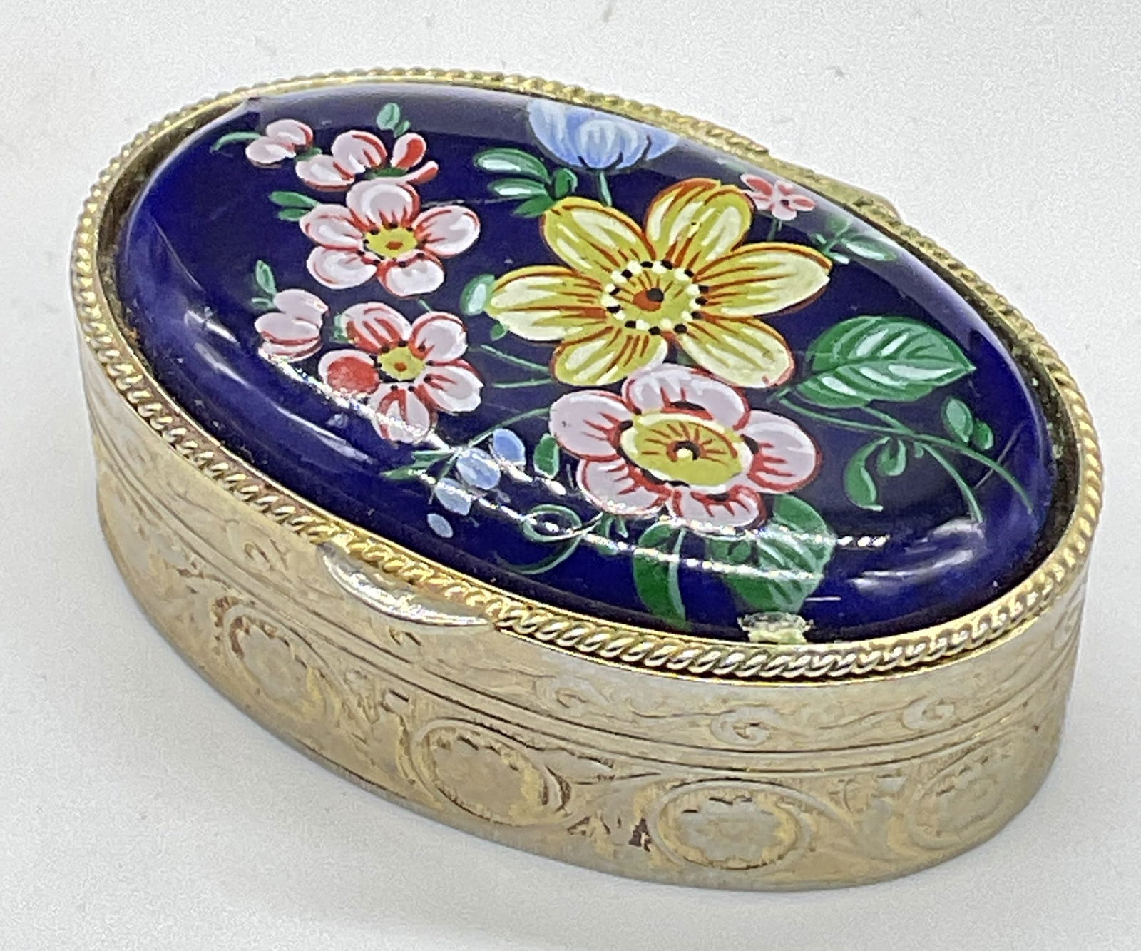 Vintage Floral Motif Metal Hinged Lid Pill Box: Vintage Floral Motif Metal Hinged Lid Pill Box / Trinket Box. Crafted primarily from metal. Cobalt blue hued costume cabochon to the lid features multicolored florals. Oval form. Measures approx. 2W x