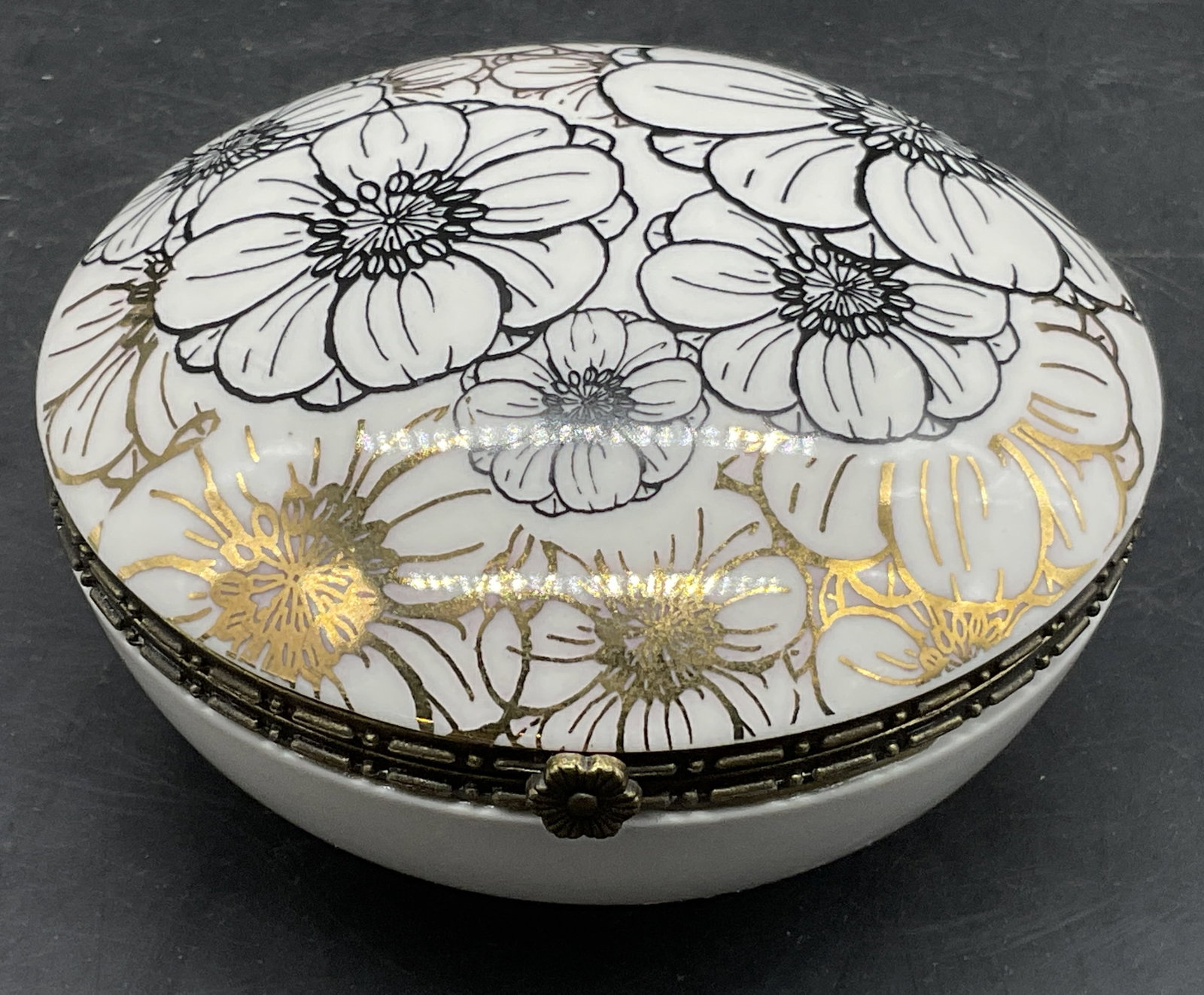 Round Porcelain Floral Motif Hinge Lid Trinket Box: Round Porcelain Floral Motif Hinged Lid Trinket Box. Primarily white with black and gilt florals to the lid. The lid opens with metal hinge hardware. Measures approx. 1.5H x 3.5W in. Discoloration and