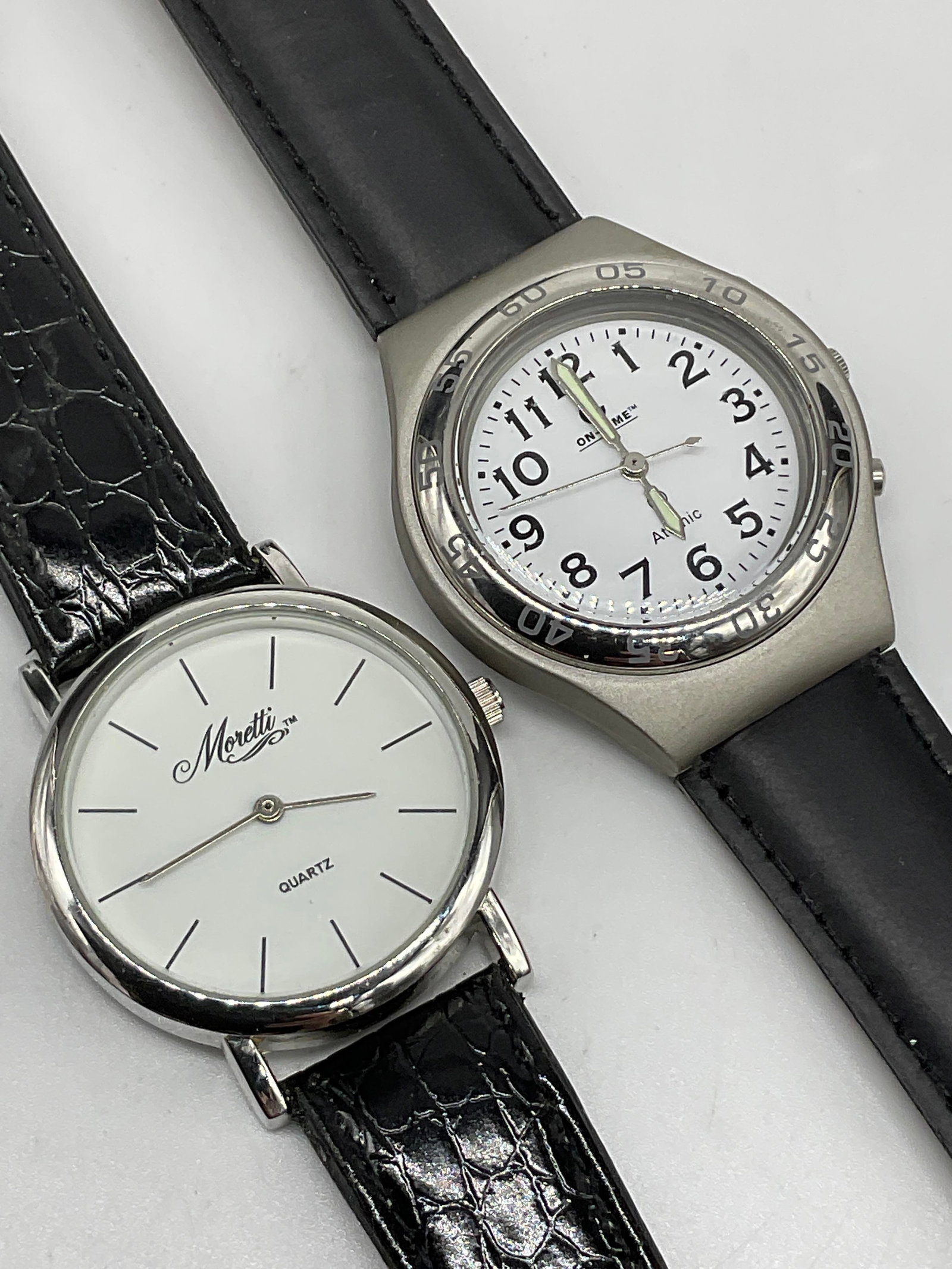 ON TIME & MORETTI Black Leather Watches: ON TIME & MORETTI Black Leather Watches. Both watches feature genuine leather bands and white hued watch faces. Largest watch measures approx. 9.75 inches in length. Surface wear consistent with age, 