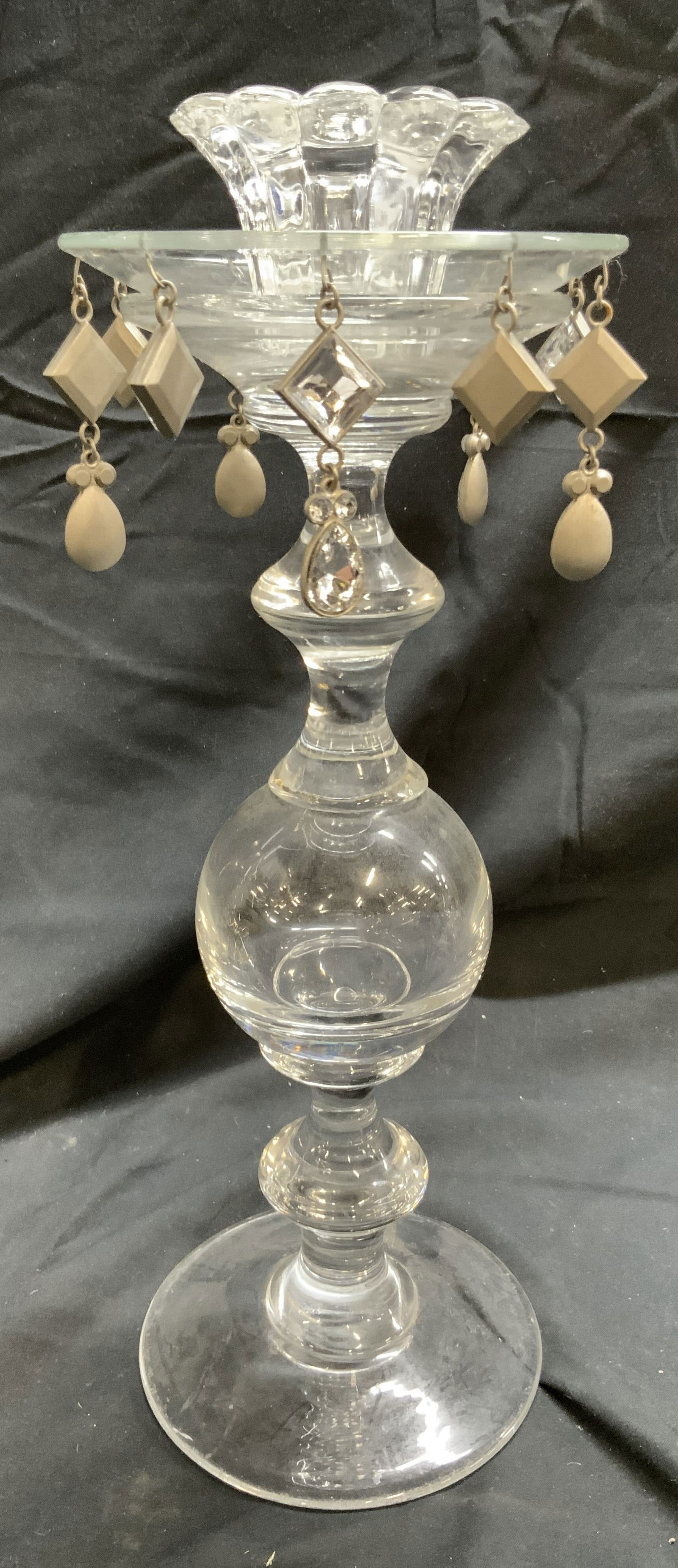 Vintage Glass Pillar Candlestick W Ornaments: Candlestick is glass. Piece has pillar form. Candlestick has hanging geometric ornaments. Base has circular form. Top of piece sits on rest of structure (not attached). Measures approximately 13 inche