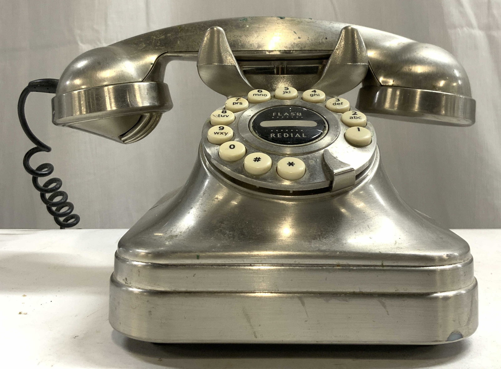 GRAND PHONE Flash Redial Brushed Metal Telephone: Underside reads GRAND PHONE. Piece is silver toned, appears to be brushed metal. Phone has circular form dial pad with flash redial button in center. Phone measures approximately 9.5 inches wide 5.5 i